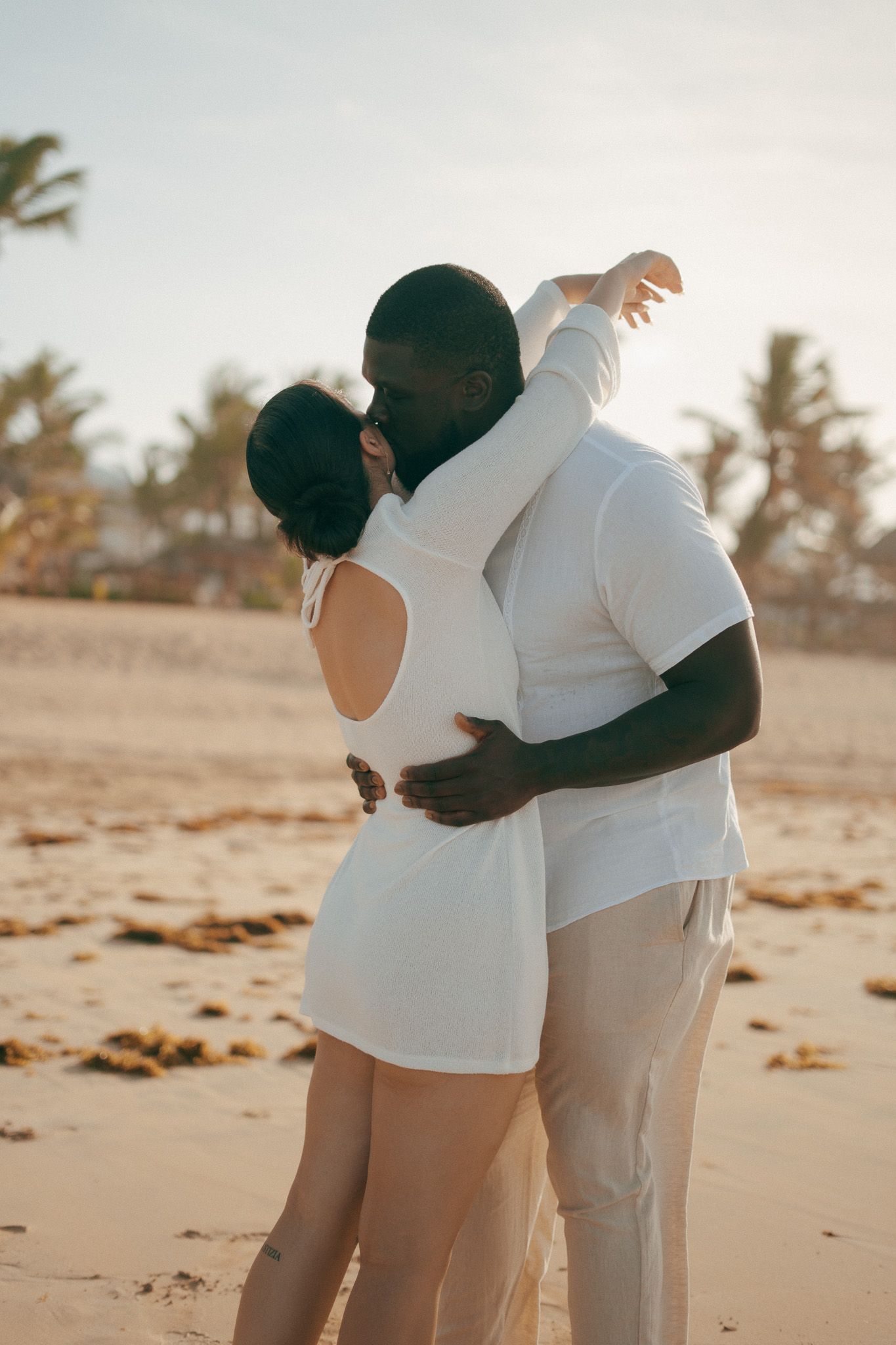 Couple in white outfits embracing and kissing on a sandy beach at golden hour with palm trees in background.