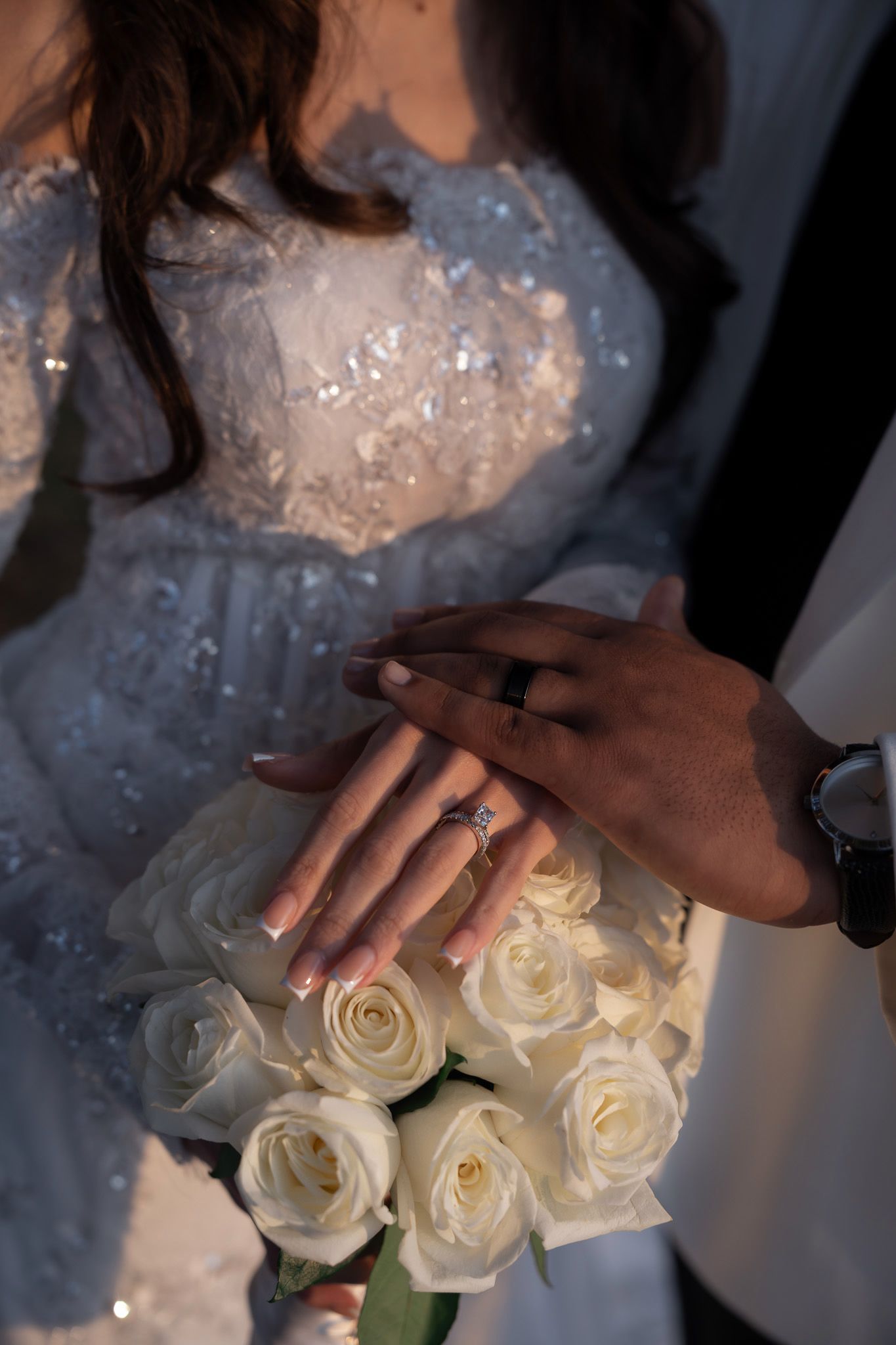Close-up of a bride holding white roses while wearing a beaded wedding dress as hands exchange rings.