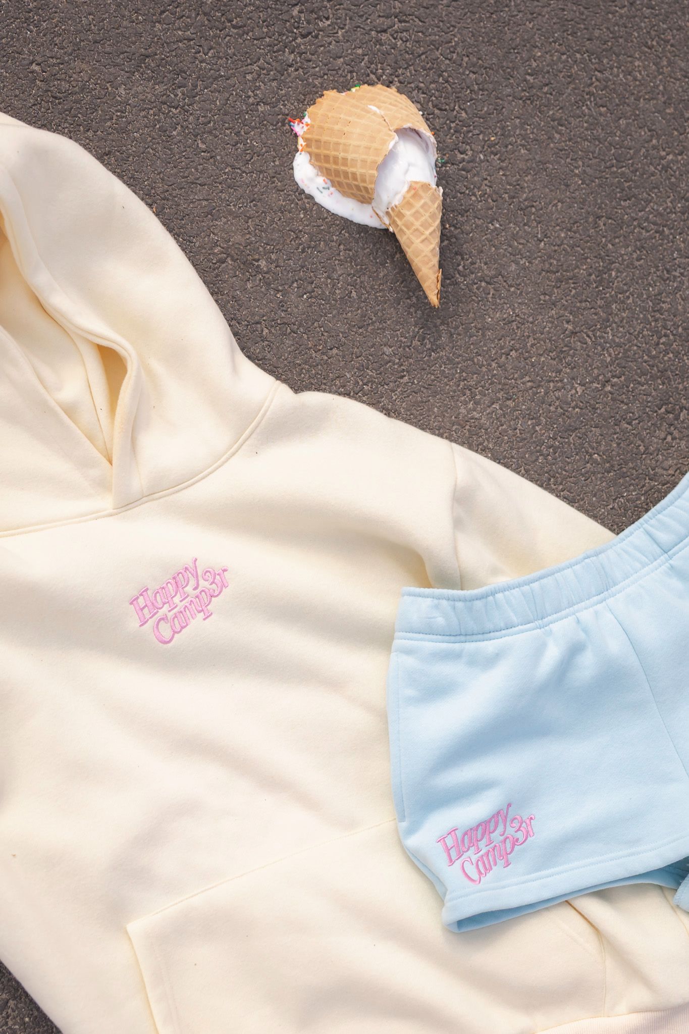 Cream hoodie and light blue shorts set with pink logo displayed flat lay beside a melting ice cream cone on pavement.