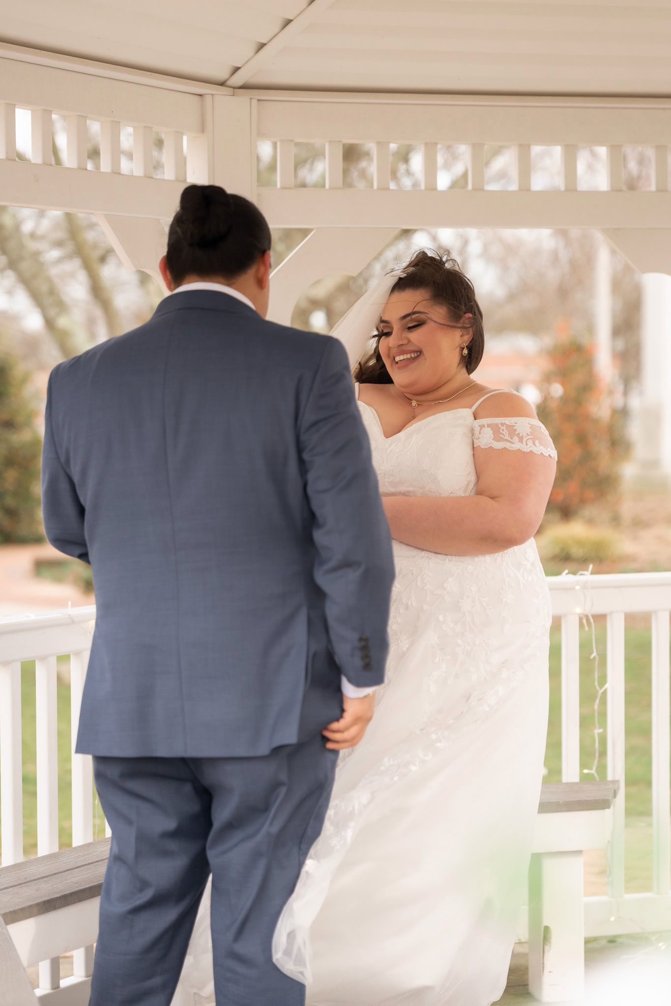 Bride in lace off-shoulder gown smiles during first look with groom in blue suit under white gazebo.
