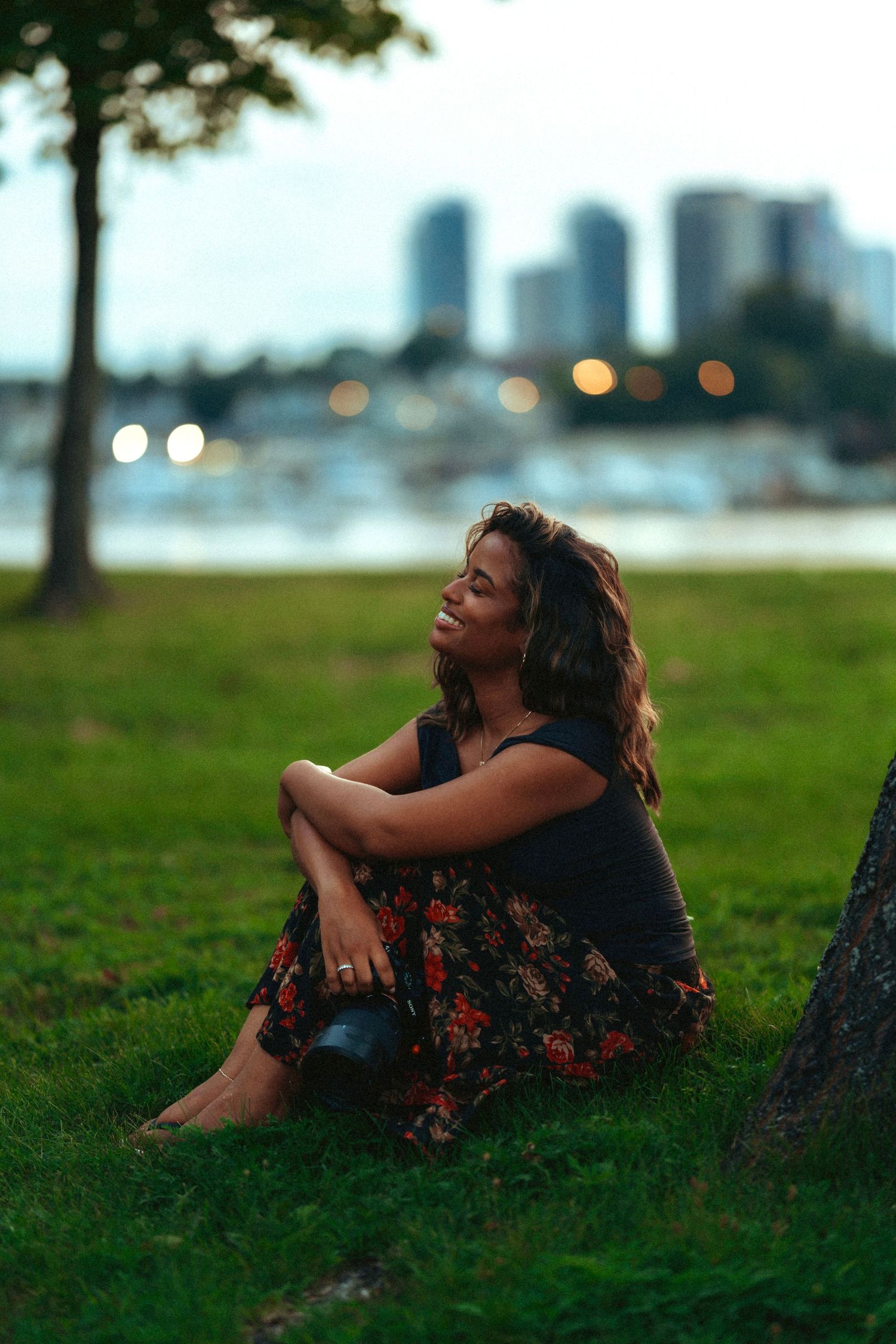Woman in floral skirt sitting barefoot on grass by waterfront park with city skyline bokeh lights at dusk.
