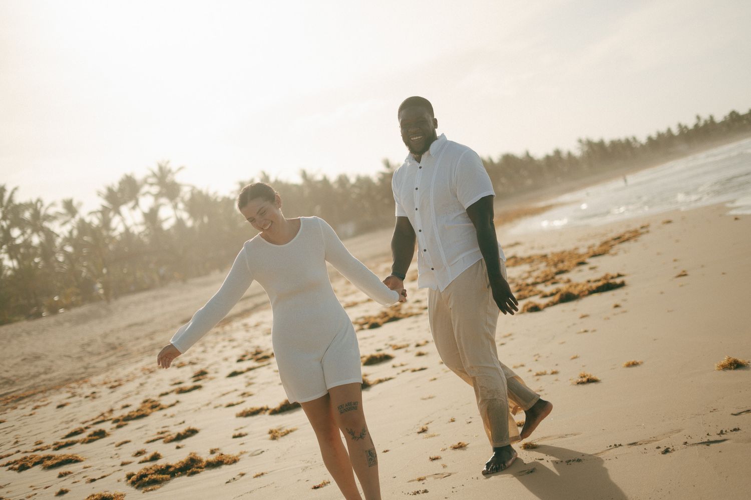 Couple holding hands walking on a sunny tropical beach, woman in white dress and man in casual attire.