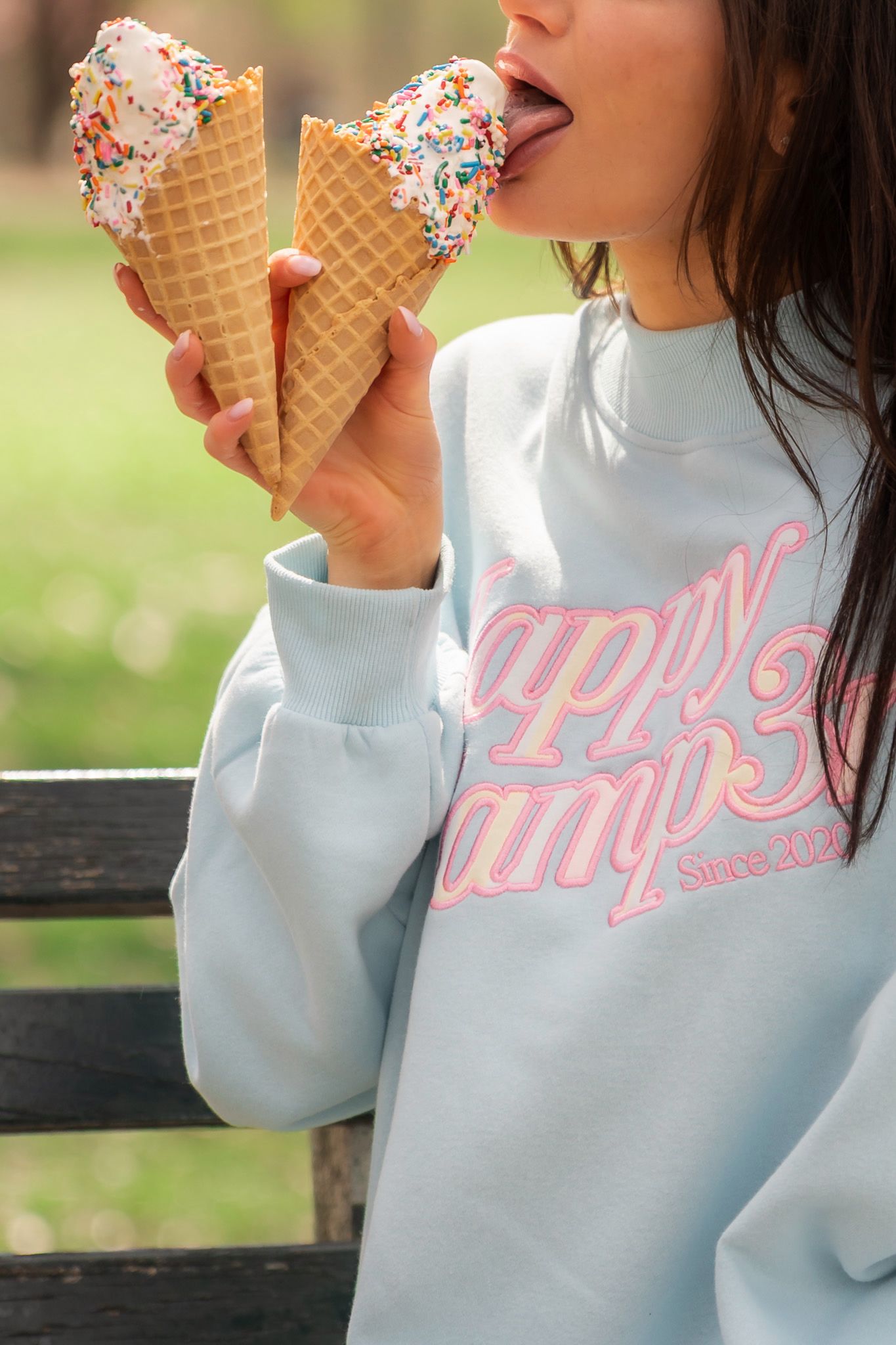 Woman in light blue Happy Camper crewneck sweatshirt licks two sprinkle ice cream cones on bench.
