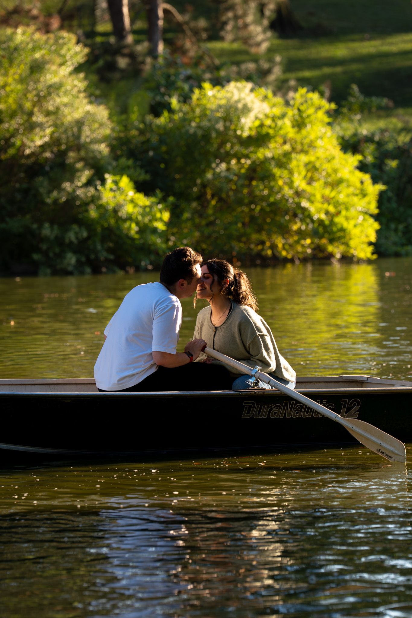 Two people sharing an intimate moment while rowing on a lake with golden sunlit trees in background.