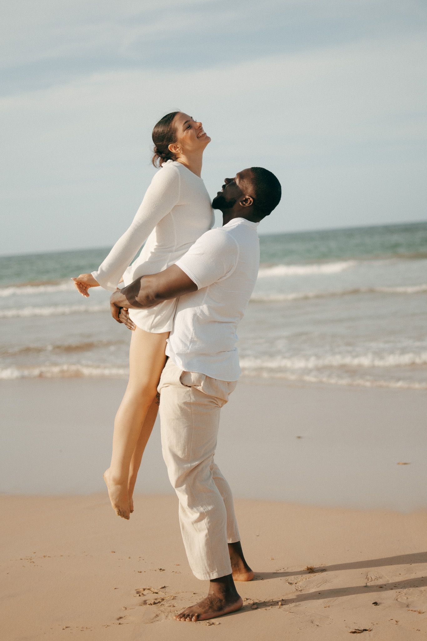 Couple in white outfits on a sandy beach, man lifting woman as ocean waves crash behind them.