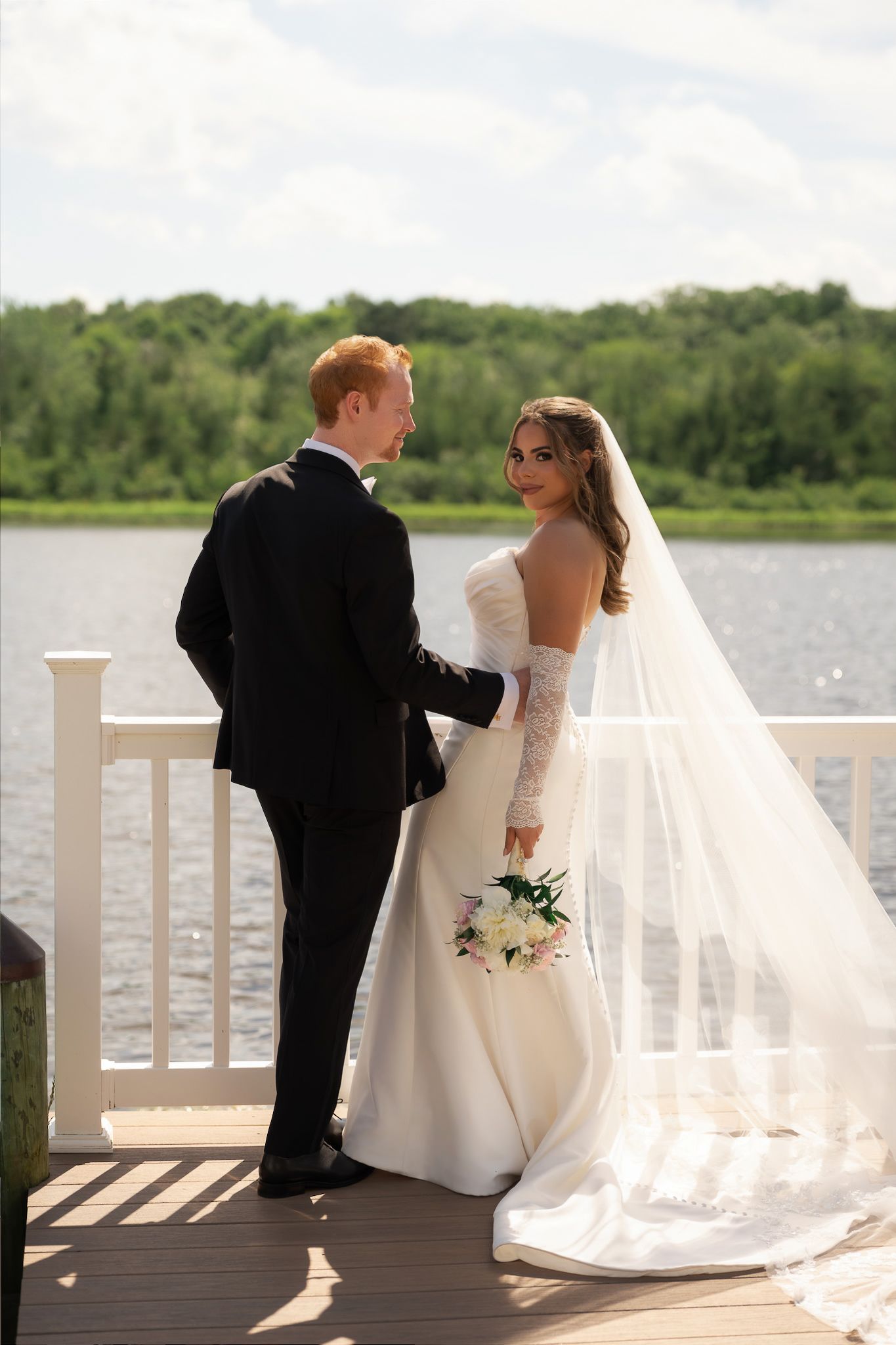 Bride and groom standing on a waterfront dock, bride holding a bouquet in a white gown with veil, groom in black suit.