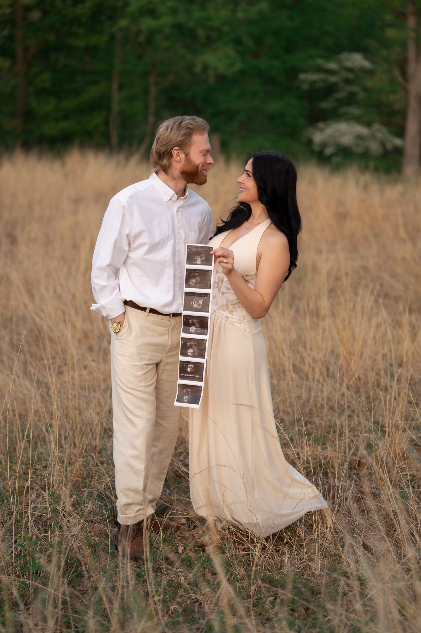 Couple standing in golden field, woman in beige dress holding ultrasound photos, man in white shirt smiling at her.