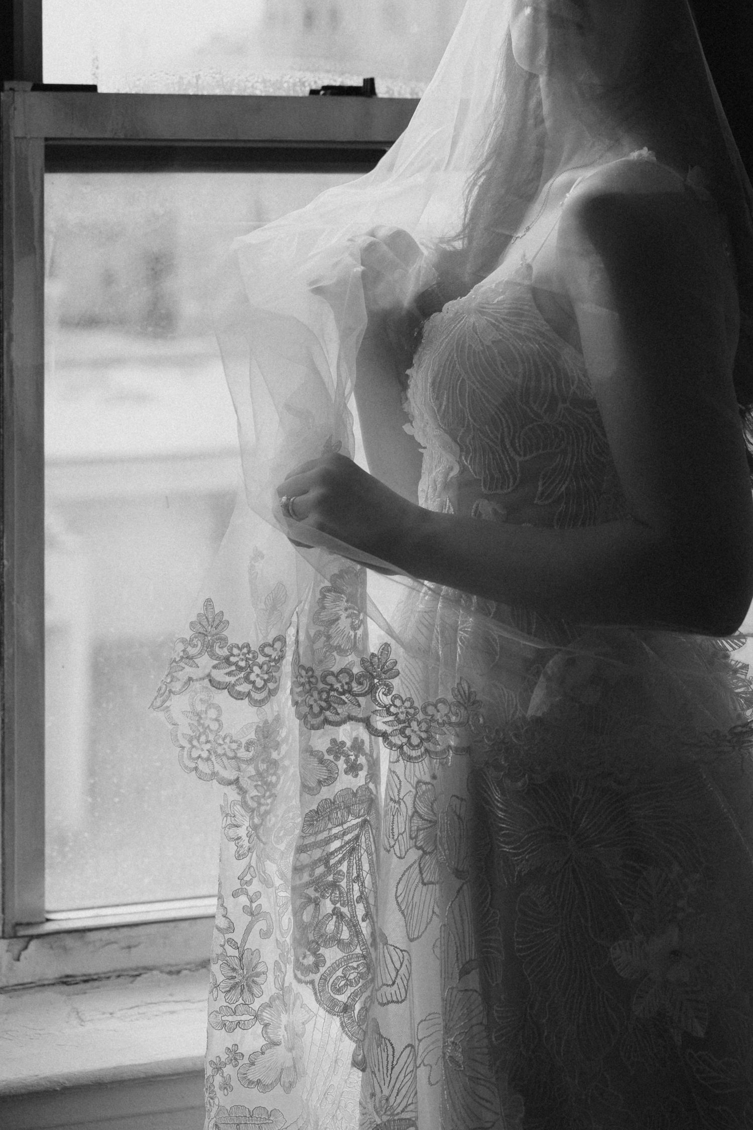 Black and white photo showing a delicate lace curtain with intricate floral patterns next to a window.