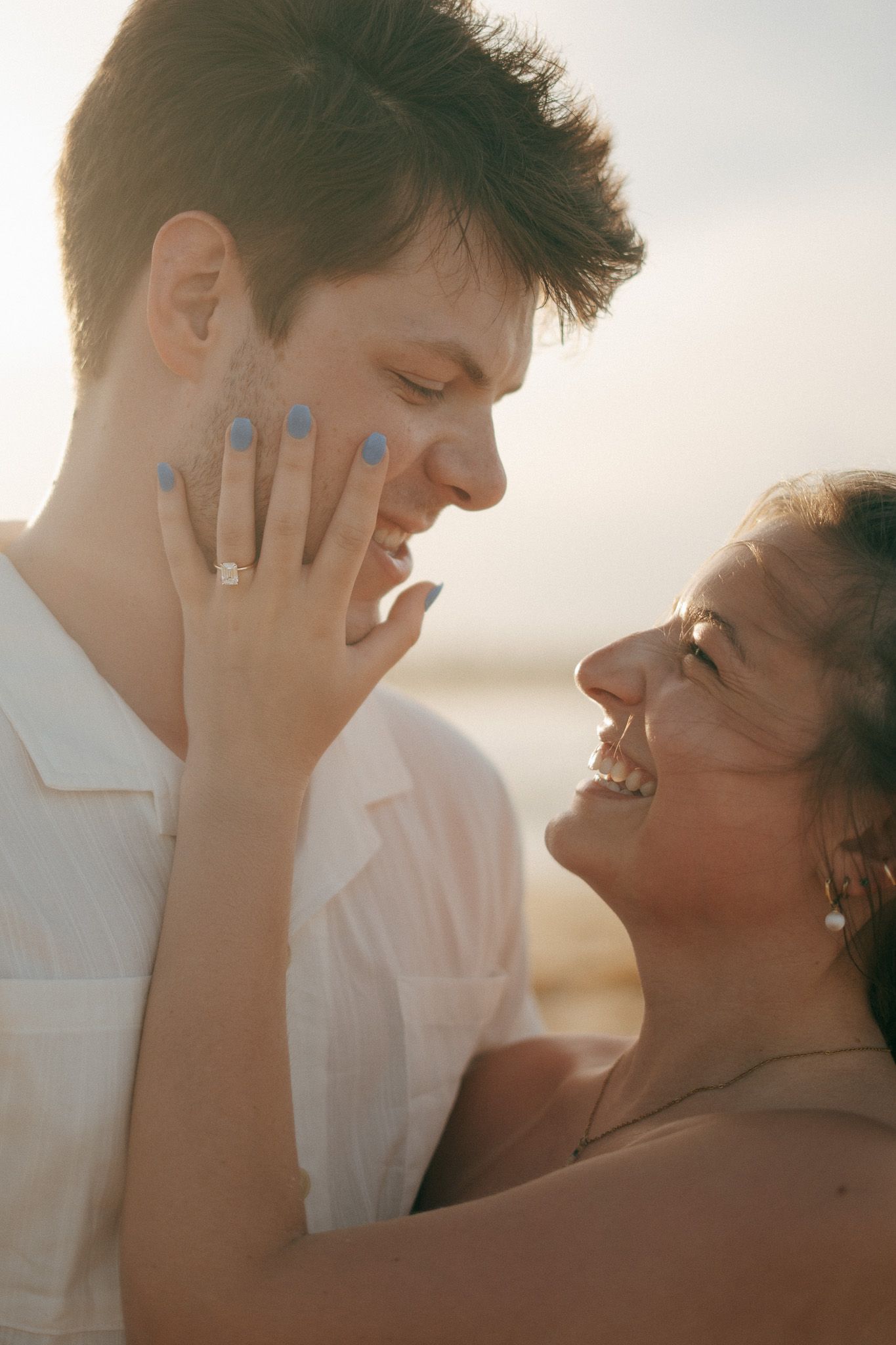 Couple sharing a romantic moment at sunset, woman showing engagement ring with blue nails, smiling joyfully.