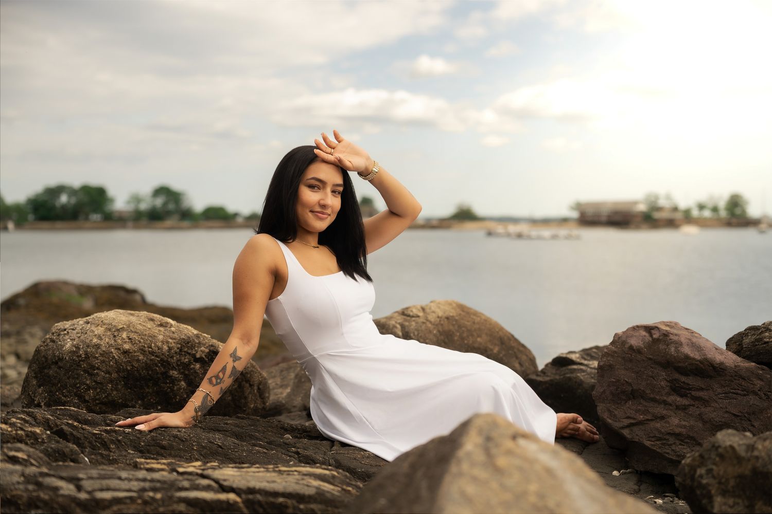 A tattooed woman in a white dress sits on rocks by the water, shielding her eyes from the sun.