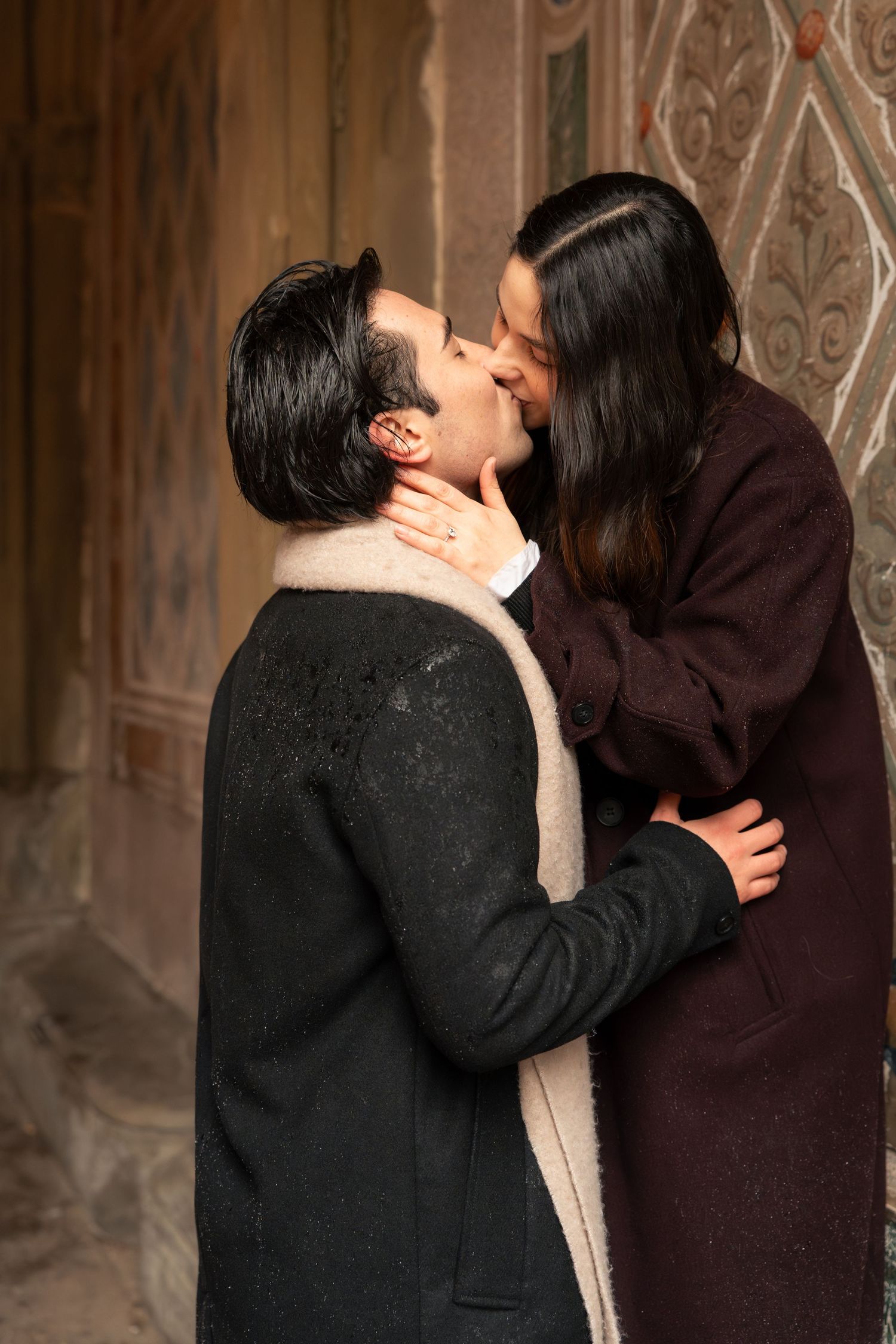 Two people in dark coats share a romantic kiss against an ornate wall background.