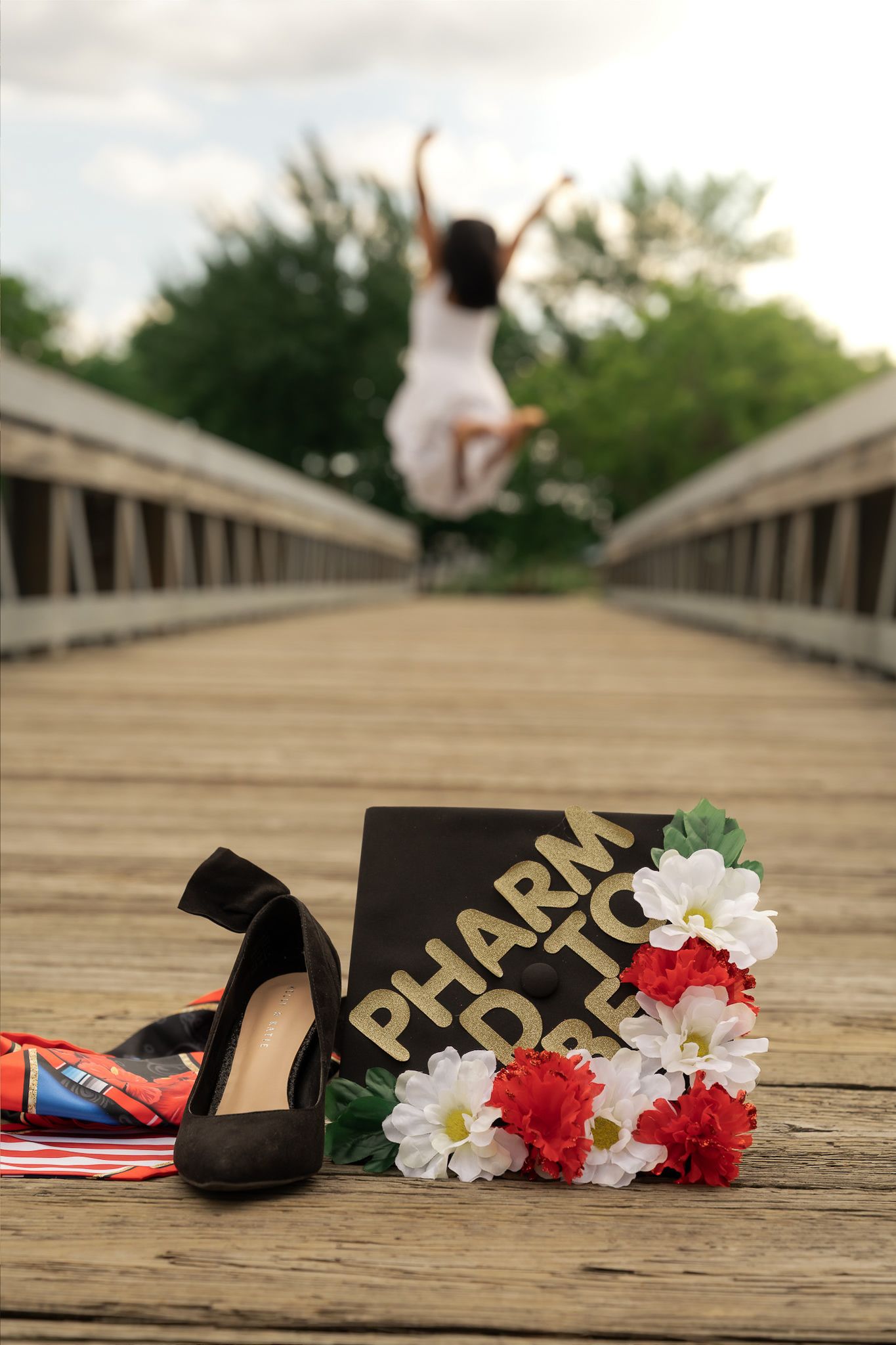 Graduate jumping for joy on bridge with PharmD cap, flowers, and heels in foreground.