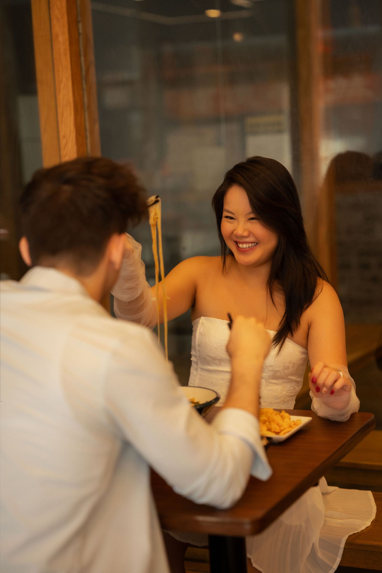 A laughing Asian woman enjoys a meal with her partner at a cozy restaurant table at night.