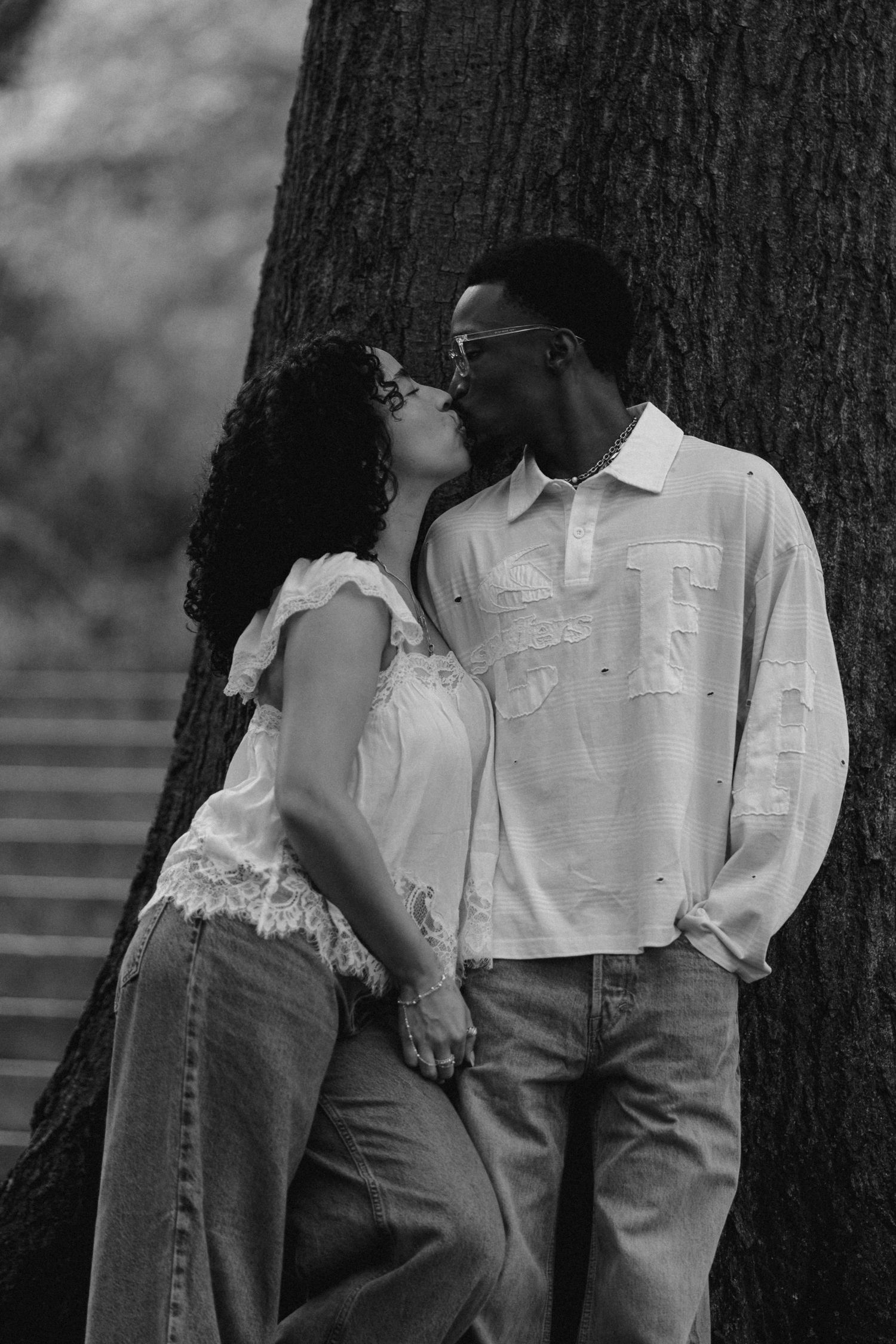 Black and white photo of a couple sharing a romantic kiss while leaning against a large tree outdoors.