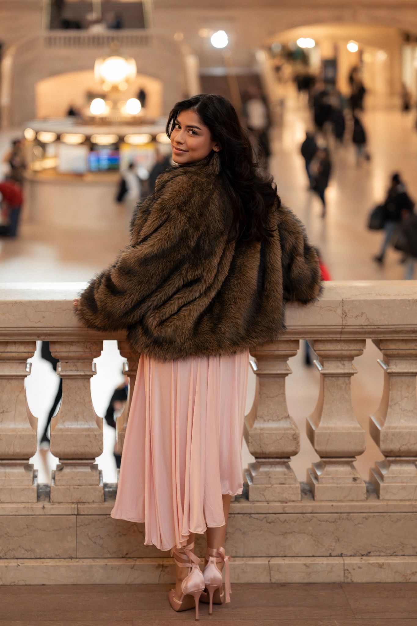 A woman in a pink dress and fur stole poses elegantly against stone balustrades in an ornate indoor setting.