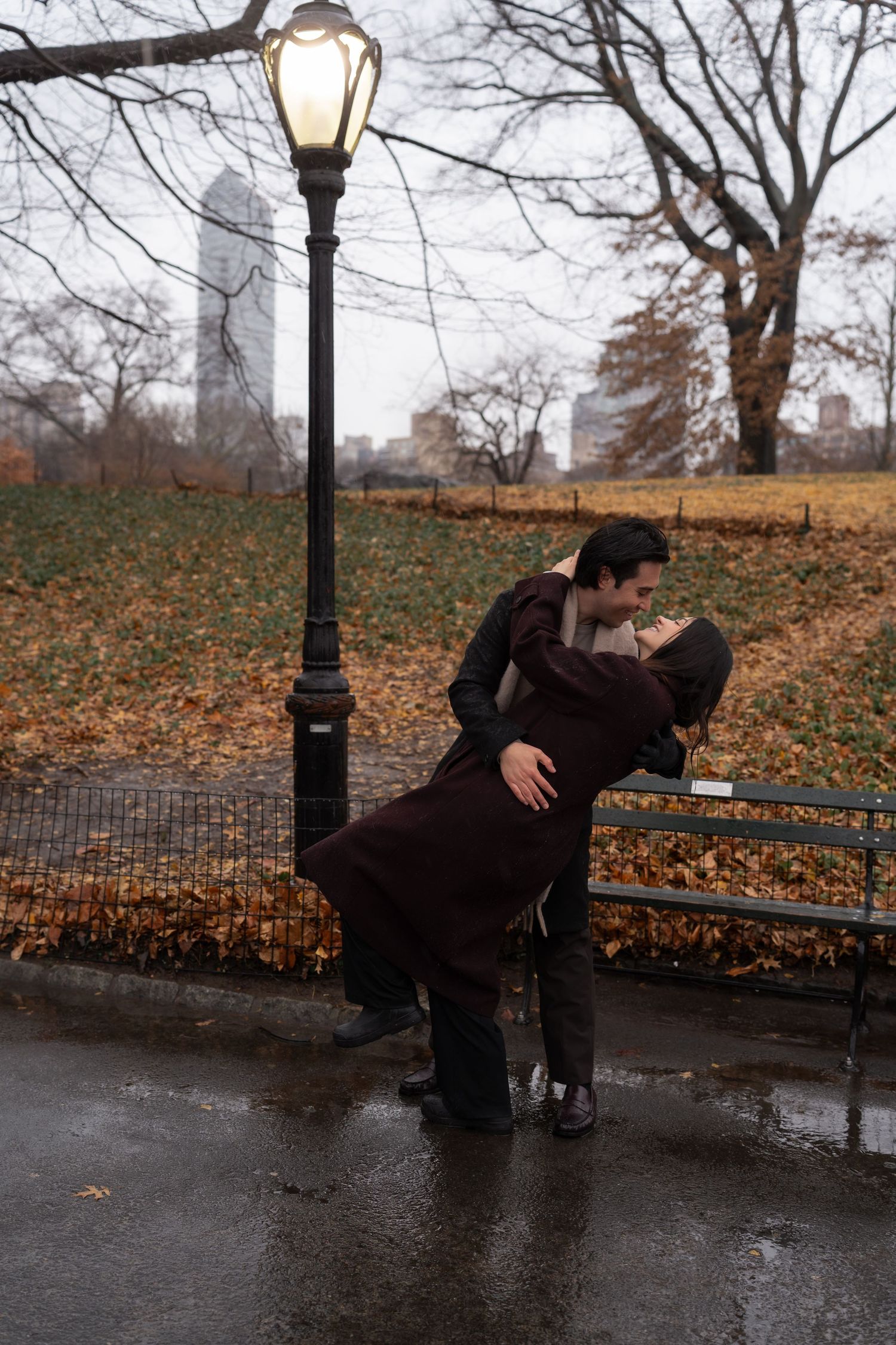 A couple embraces on a park bench beneath a lit lamppost on a rainy autumn day in Central Park.