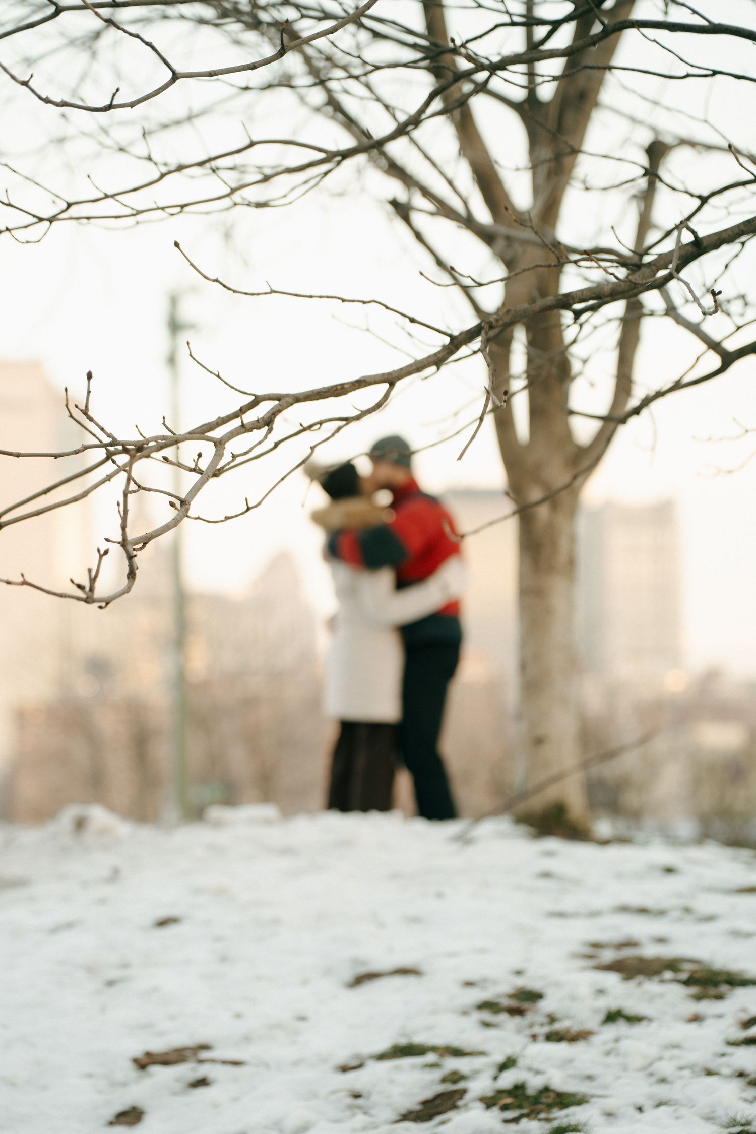 A couple embraces while standing on a snowy hill with bare winter trees and city buildings in the background.