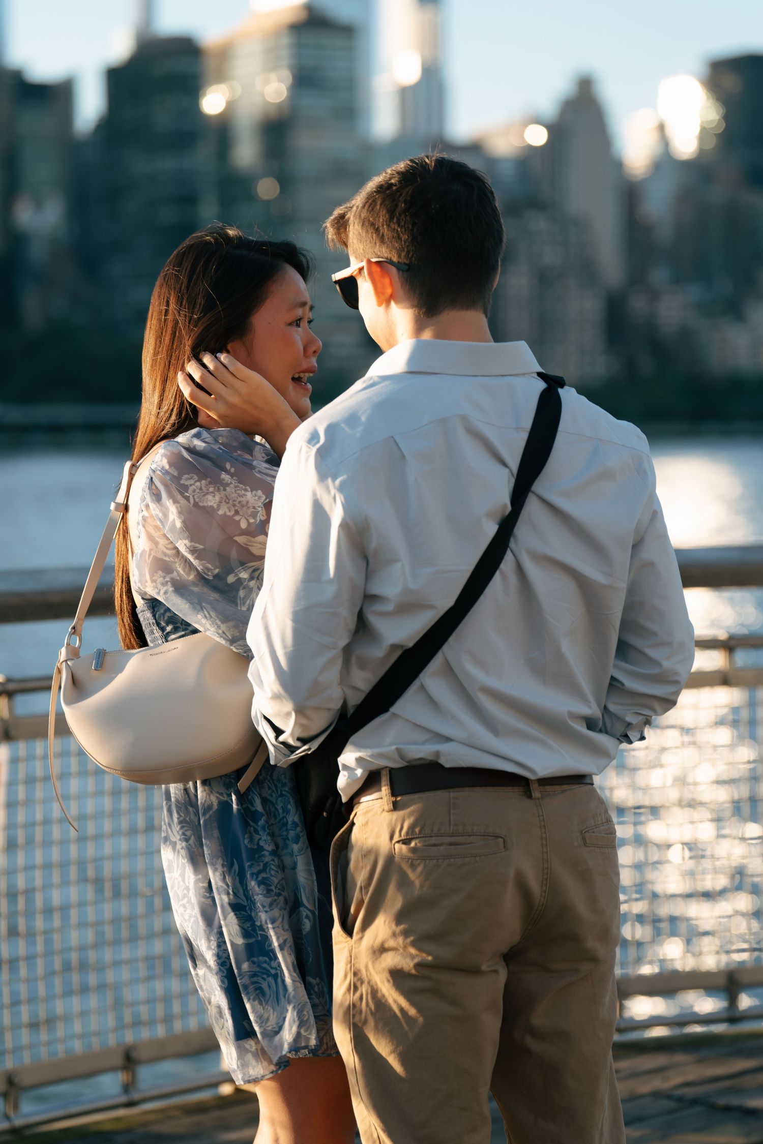 A couple shares an intimate moment at sunset along the waterfront with the city skyline glowing in the background.