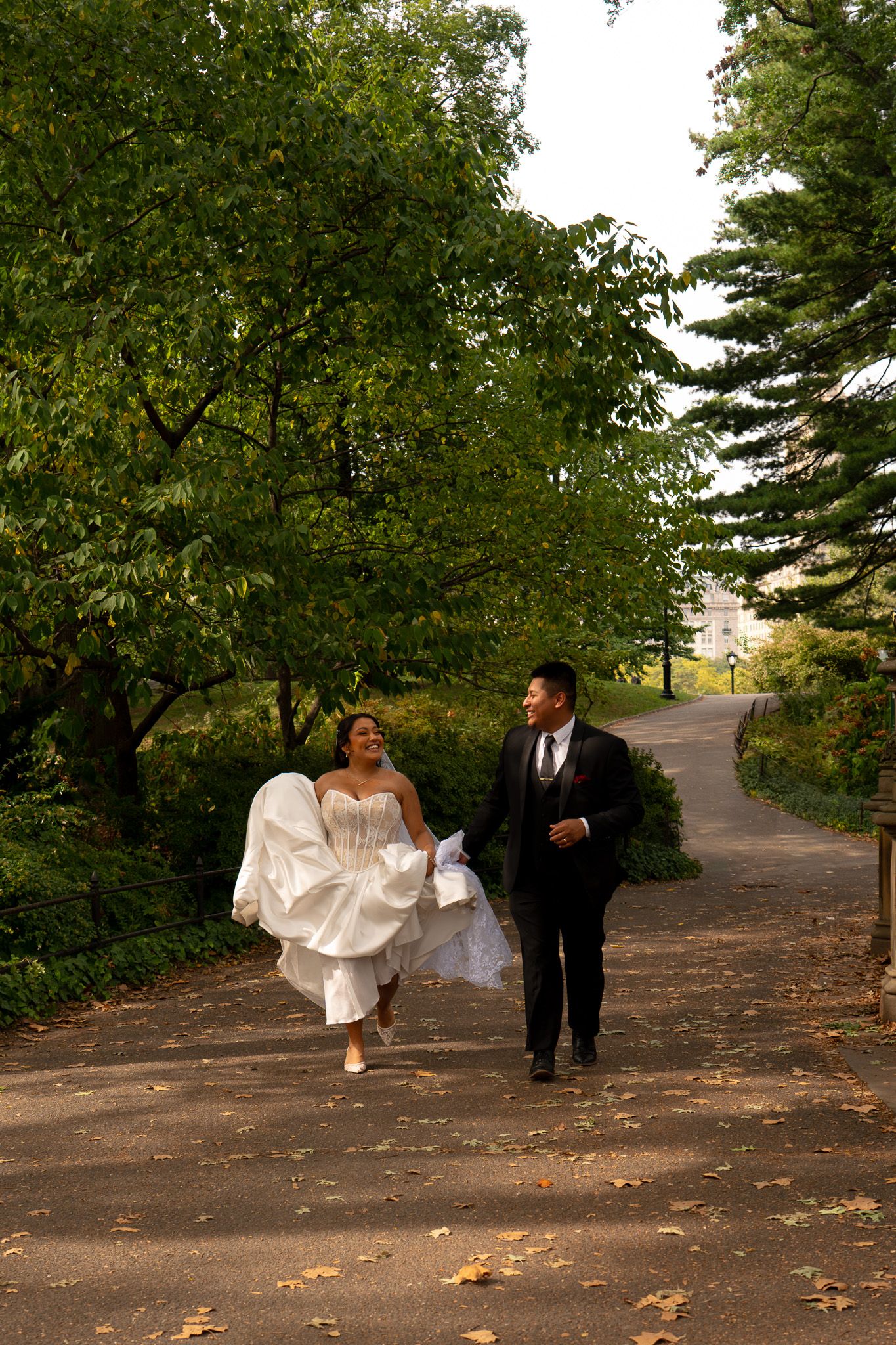 A couple walks together on a tree-lined autumn path, with the bride holding up her white dress.