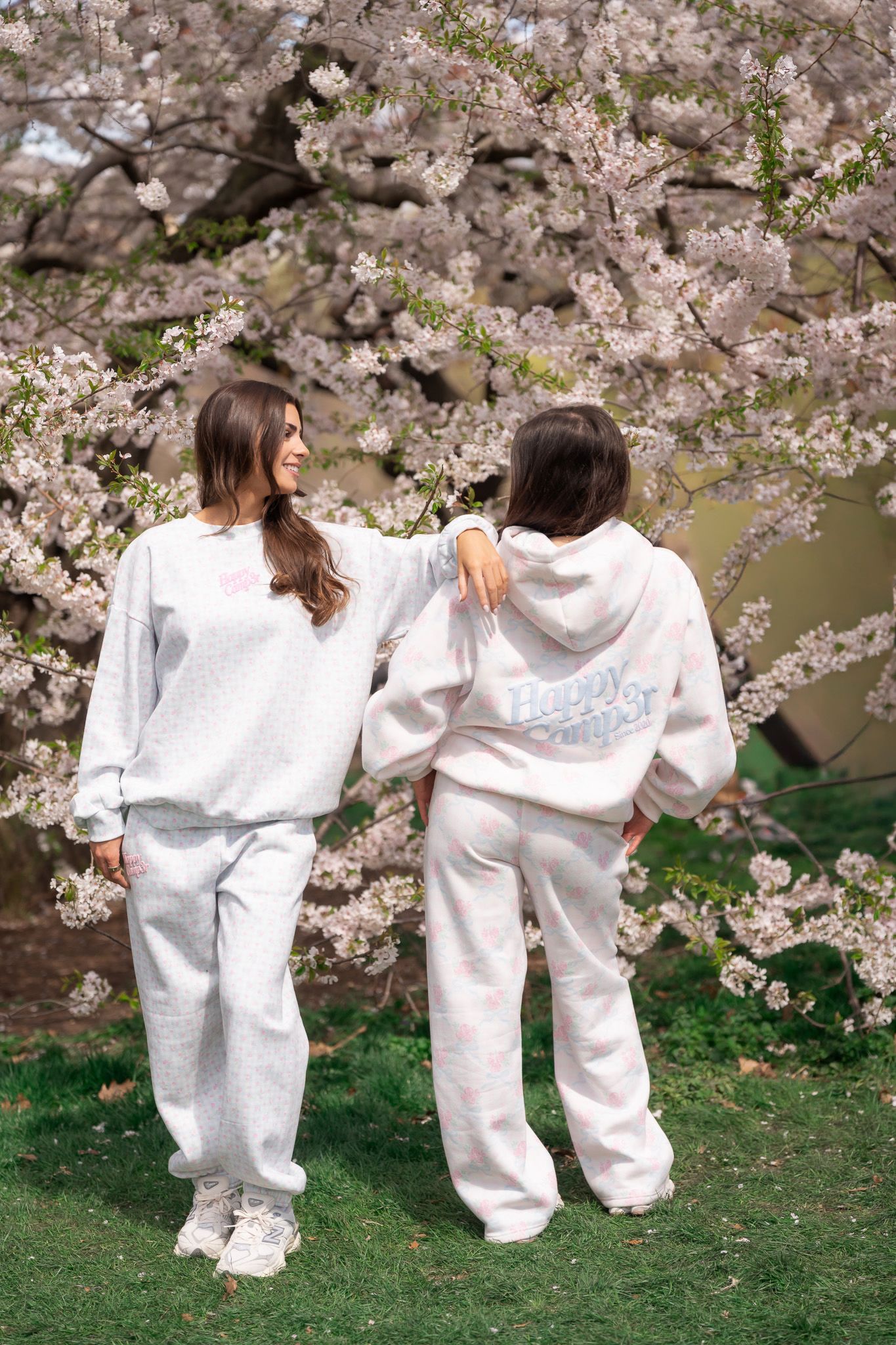 Two women in matching gray Happy Camper sweatsuits pose under blooming cherry blossom trees on green grass.