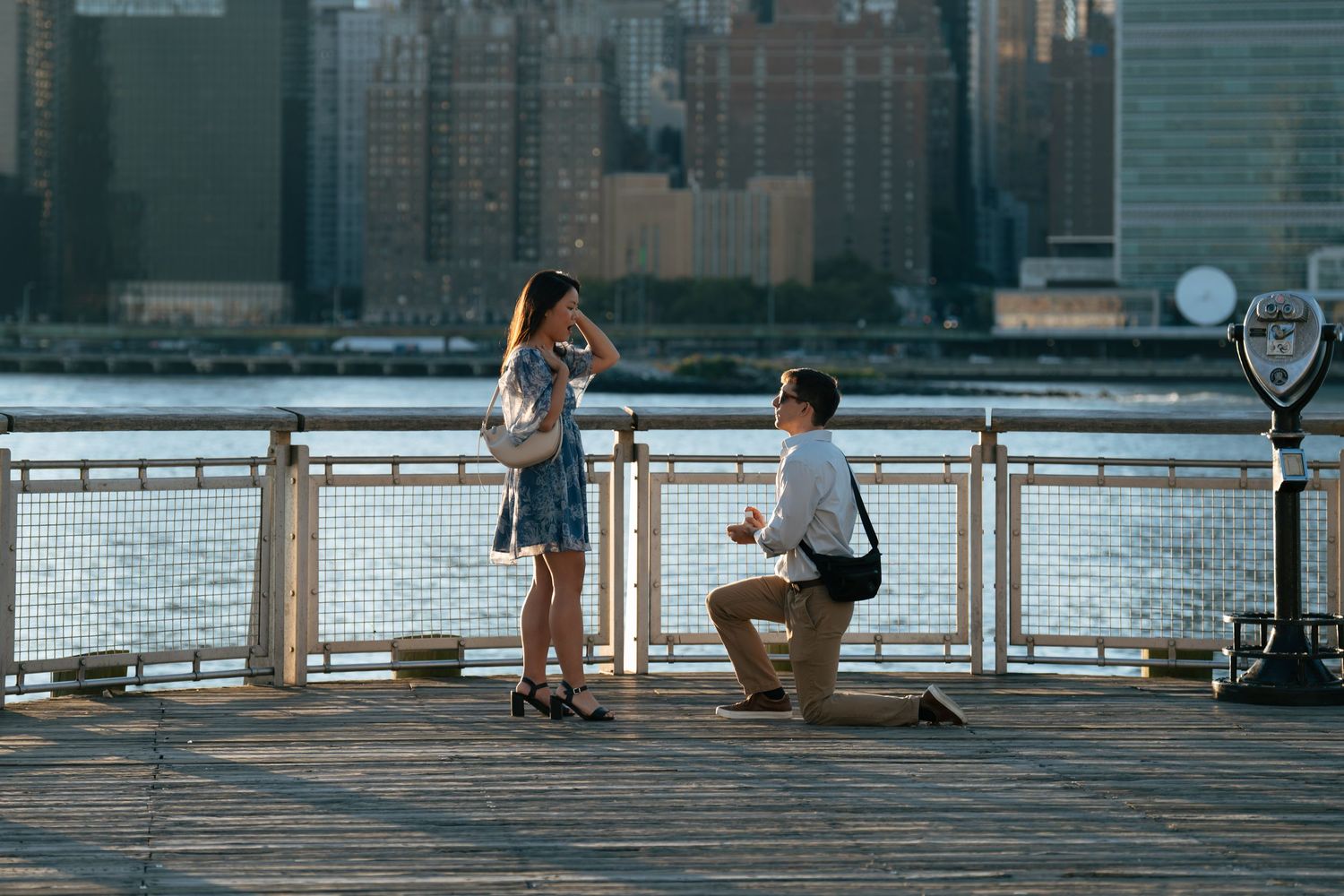 A couple shares a romantic moment during a sunset proposal on a waterfront pier with the city skyline in the background.