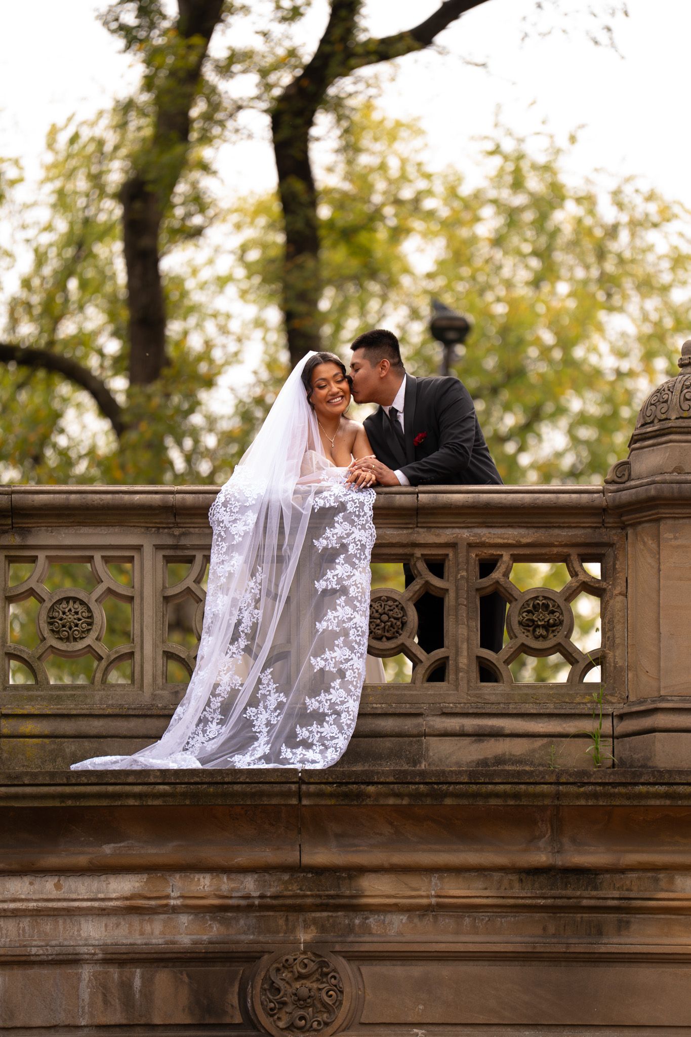A romantic wedding couple shares a tender moment on an ornate stone balcony surrounded by autumn trees.