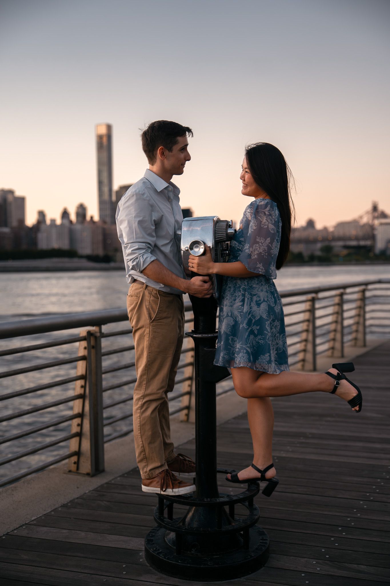 Couple playing on vintage viewfinder telescope on waterfront boardwalk at sunset.