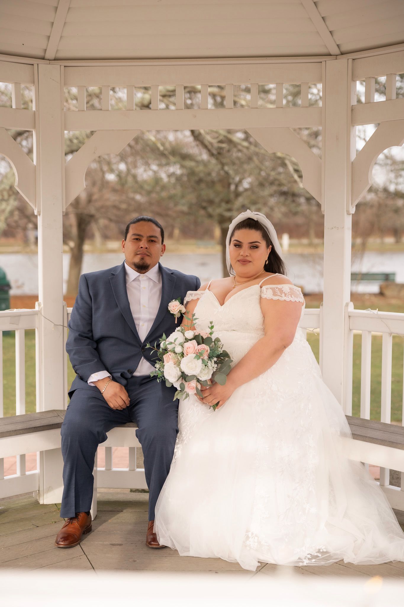 Bride and groom posing in a white gazebo, bride in lace gown holding pink bouquet, groom in gray suit.