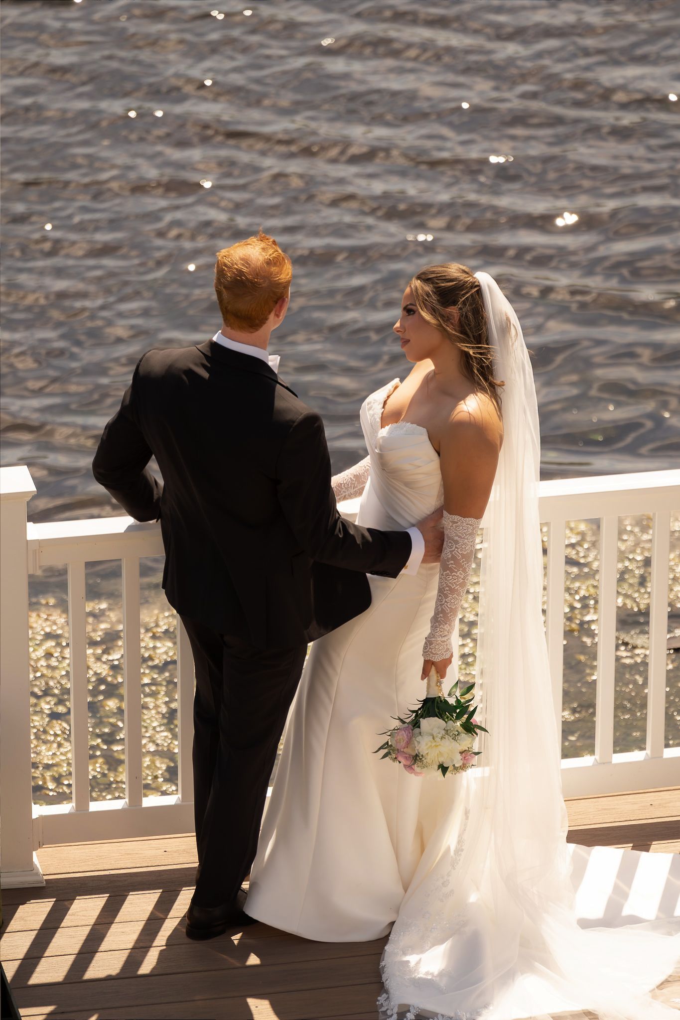 Bride and groom stand on waterfront deck, bride in white gown with veil holding bouquet, groom in black suit.