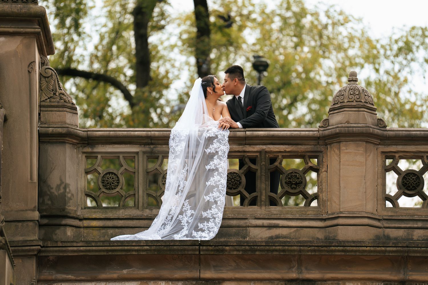 Elegant wedding couple embraces on a stone balcony surrounded by lush trees on their special day.