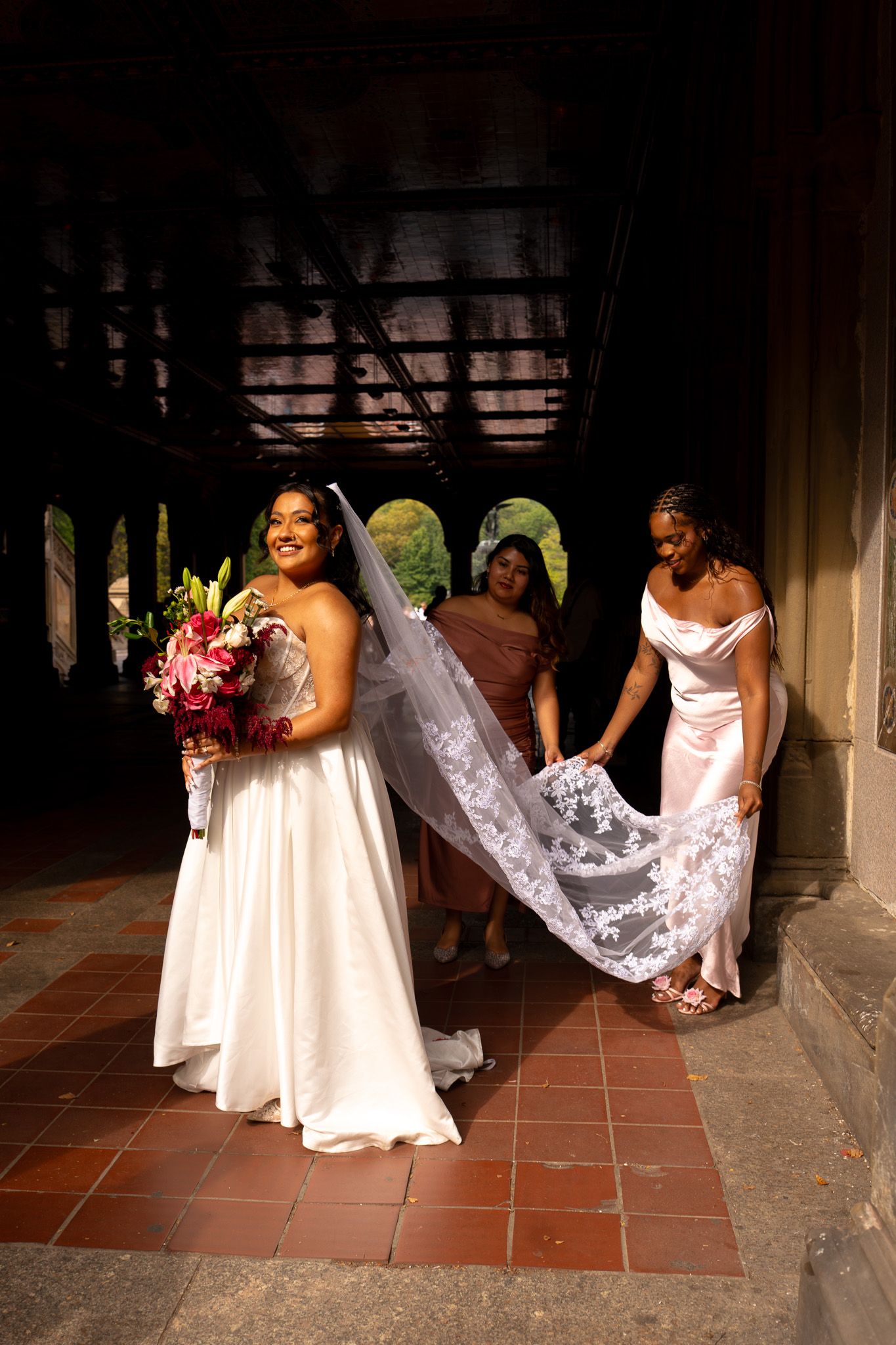 Wedding party in off-shoulder gowns walks along covered walkway with dramatic lace veil trailing behind.