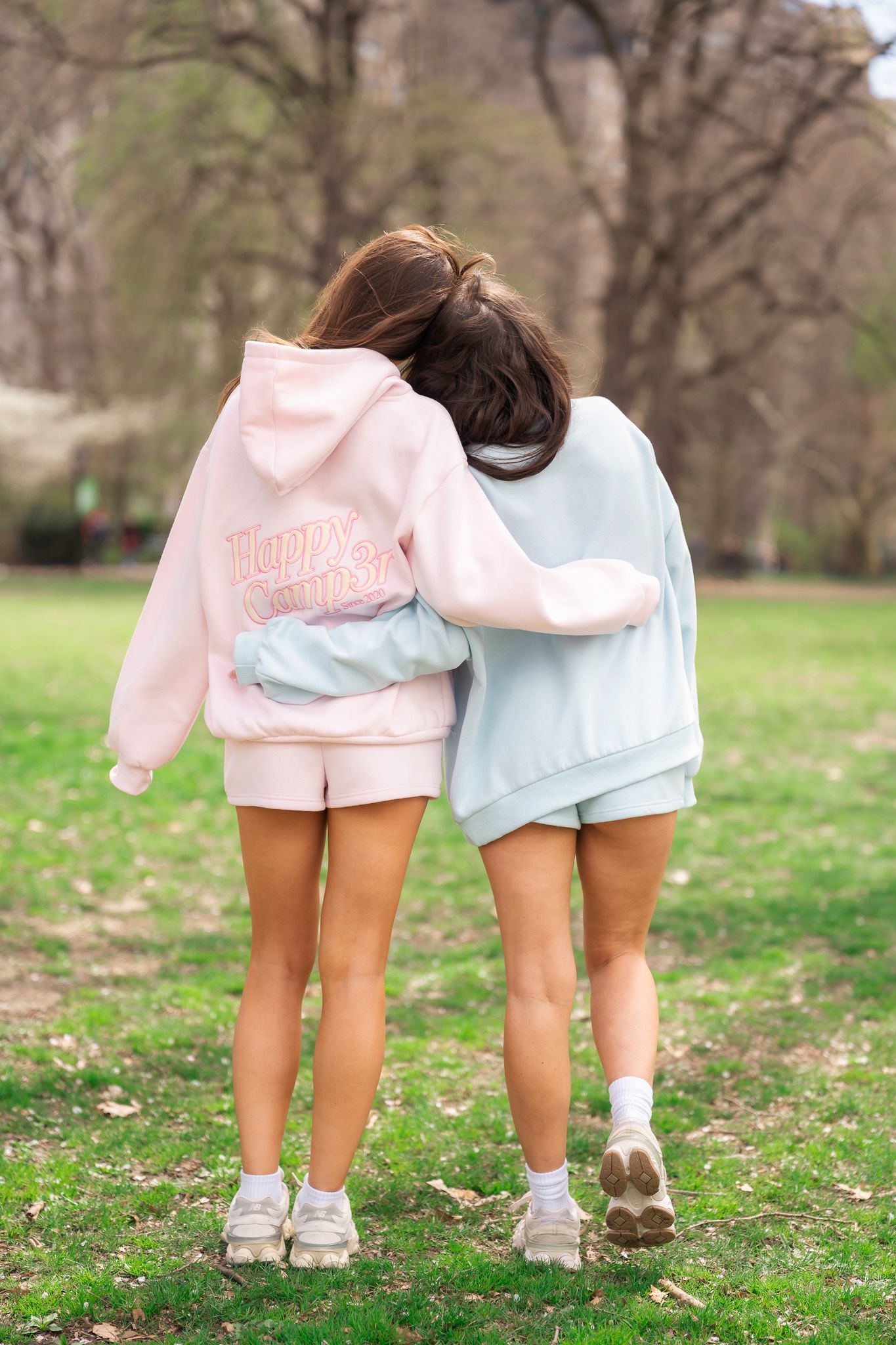Two girls in matching pastel pink and blue hoodies and shorts walking arm in arm in a park.