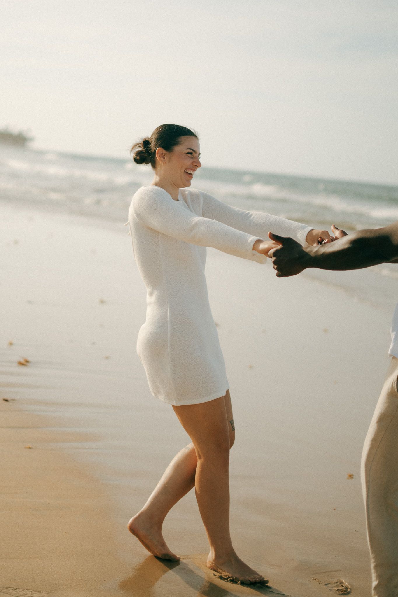 A smiling woman in a white dress holds hands with someone on a sunny beach, waves crashing in the background.