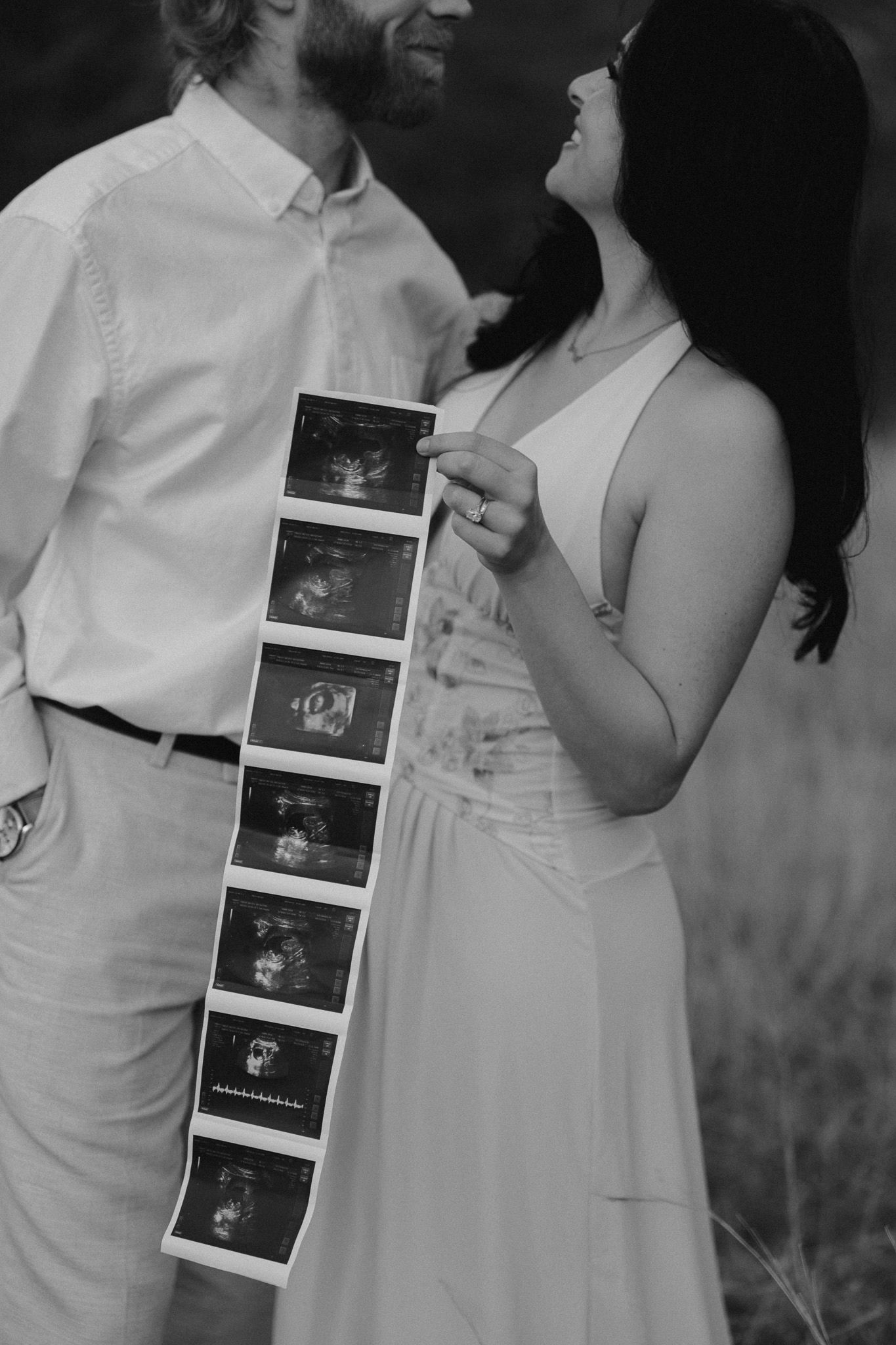 Black and white photo of a couple holding ultrasound strip, pregnancy announcement portrait outdoors.