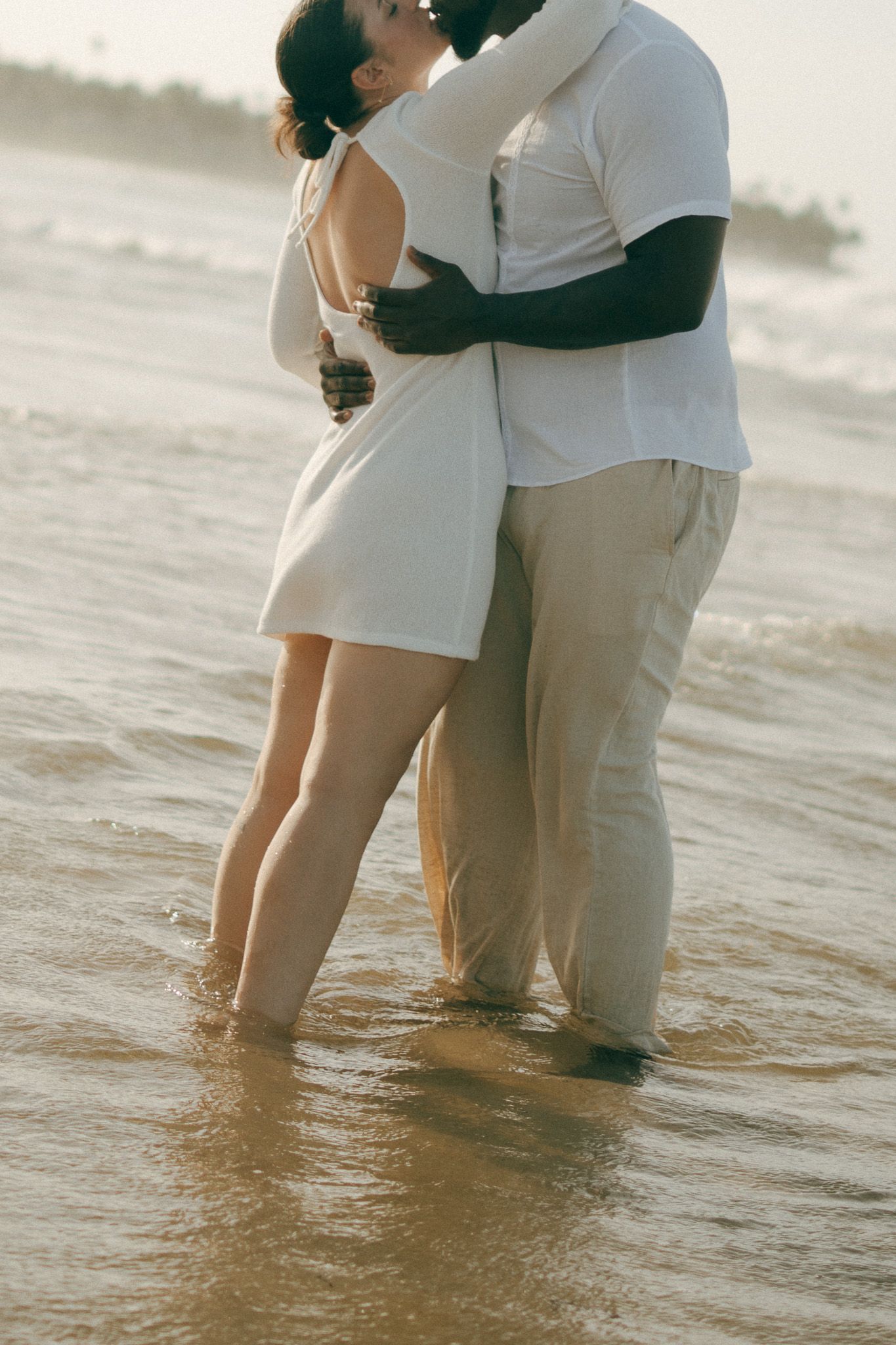 Romantic interracial couple embracing and kissing at the beach shoreline, dressed in white and beige outfits.