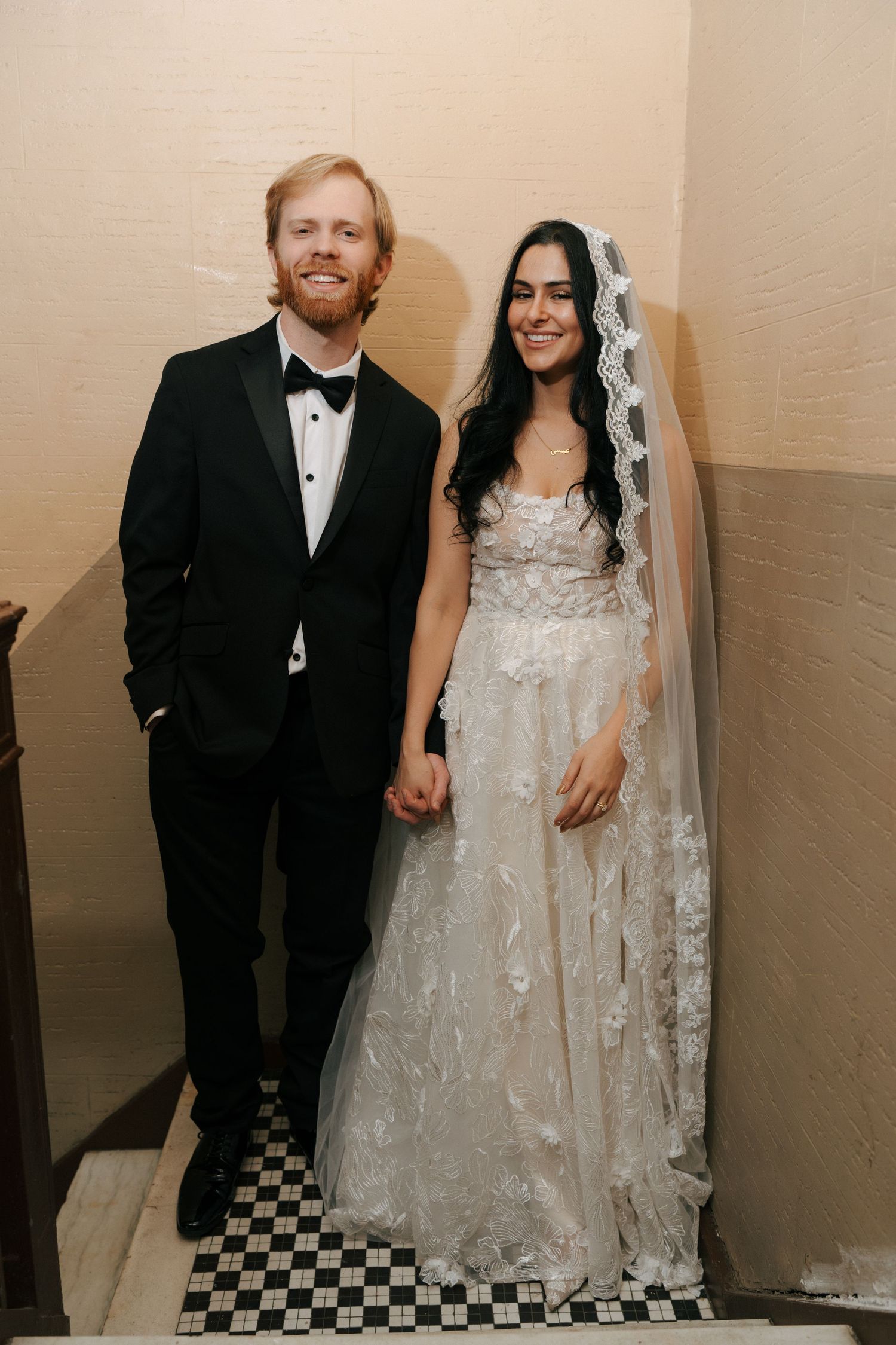 A couple poses for wedding photos in a hallway with black and white checkered floor tiles.