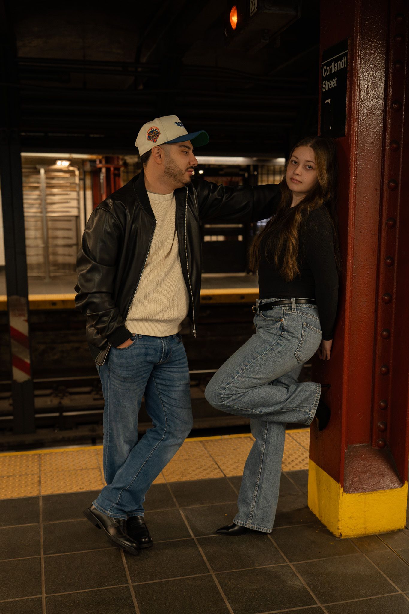 Two people lean against a subway platform column in casual denim and leather attire.