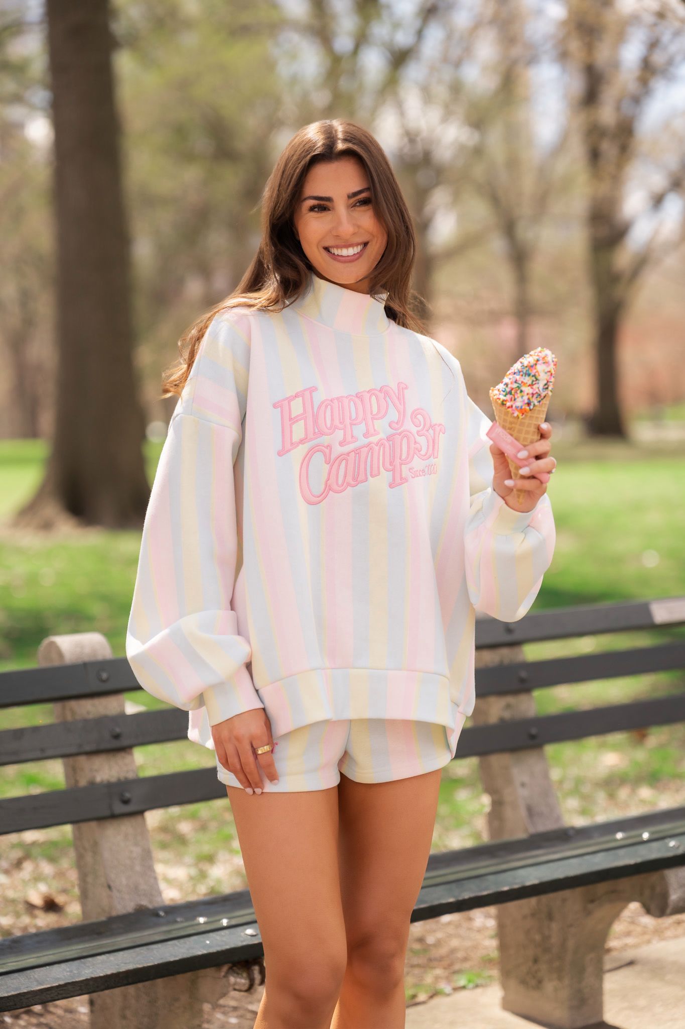 Smiling woman in pastel striped Happy Camper sweatshirt holds sprinkle ice cream cone in sunny park.