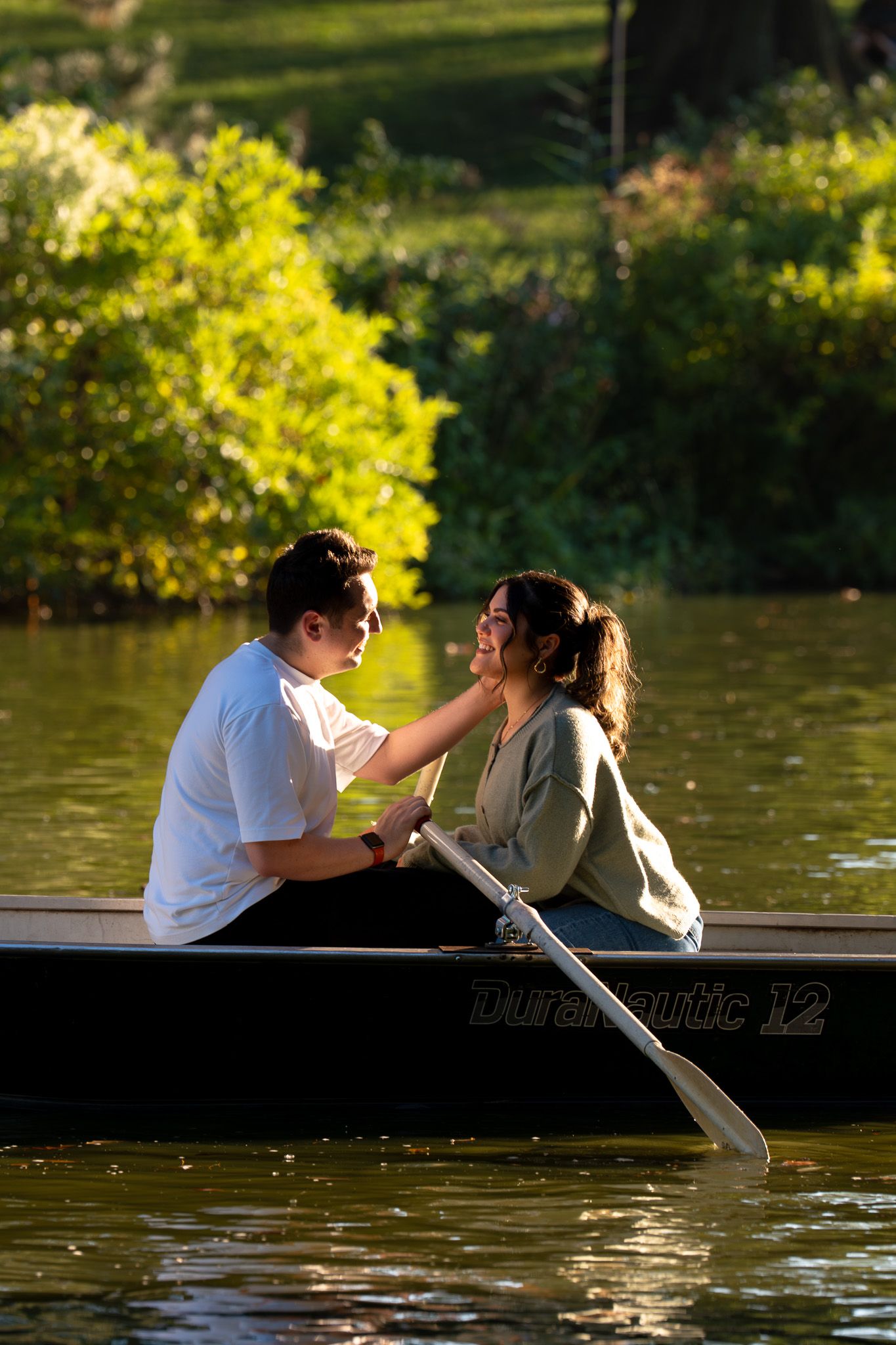 A couple shares an intimate moment while sitting in a rowboat on a lake surrounded by golden sunlit foliage.