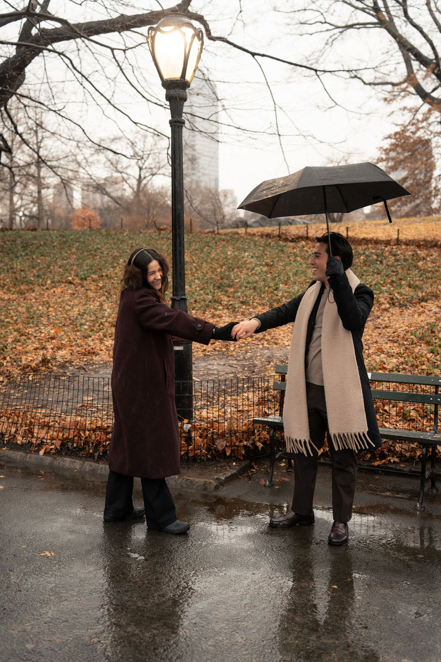 Two people sharing a romantic moment in the rain under an umbrella near a glowing lamppost in Central Park during autumn.
