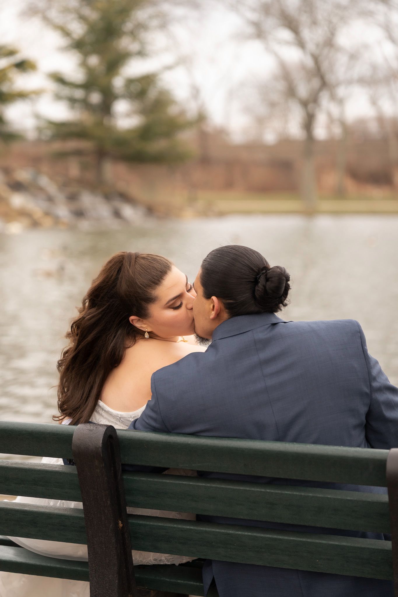 Couple sharing a romantic kiss on a green park bench beside a tranquil lake, dressed in wedding attire.