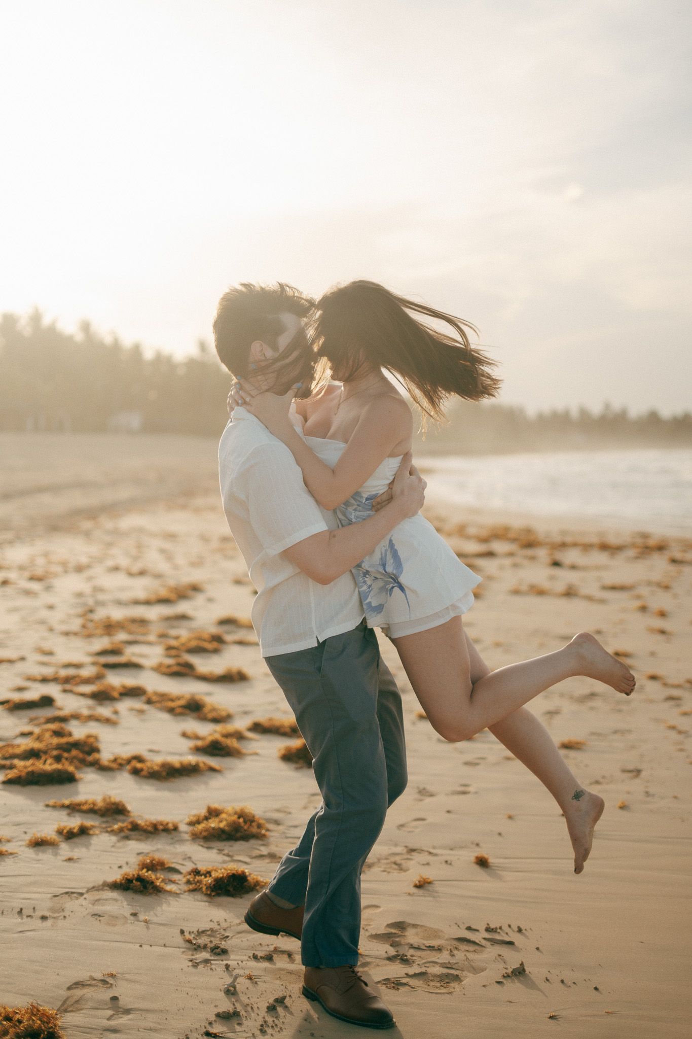 Couple sharing a romantic kiss on a sandy beach at golden sunset, man lifting woman as her hair blows in the wind.