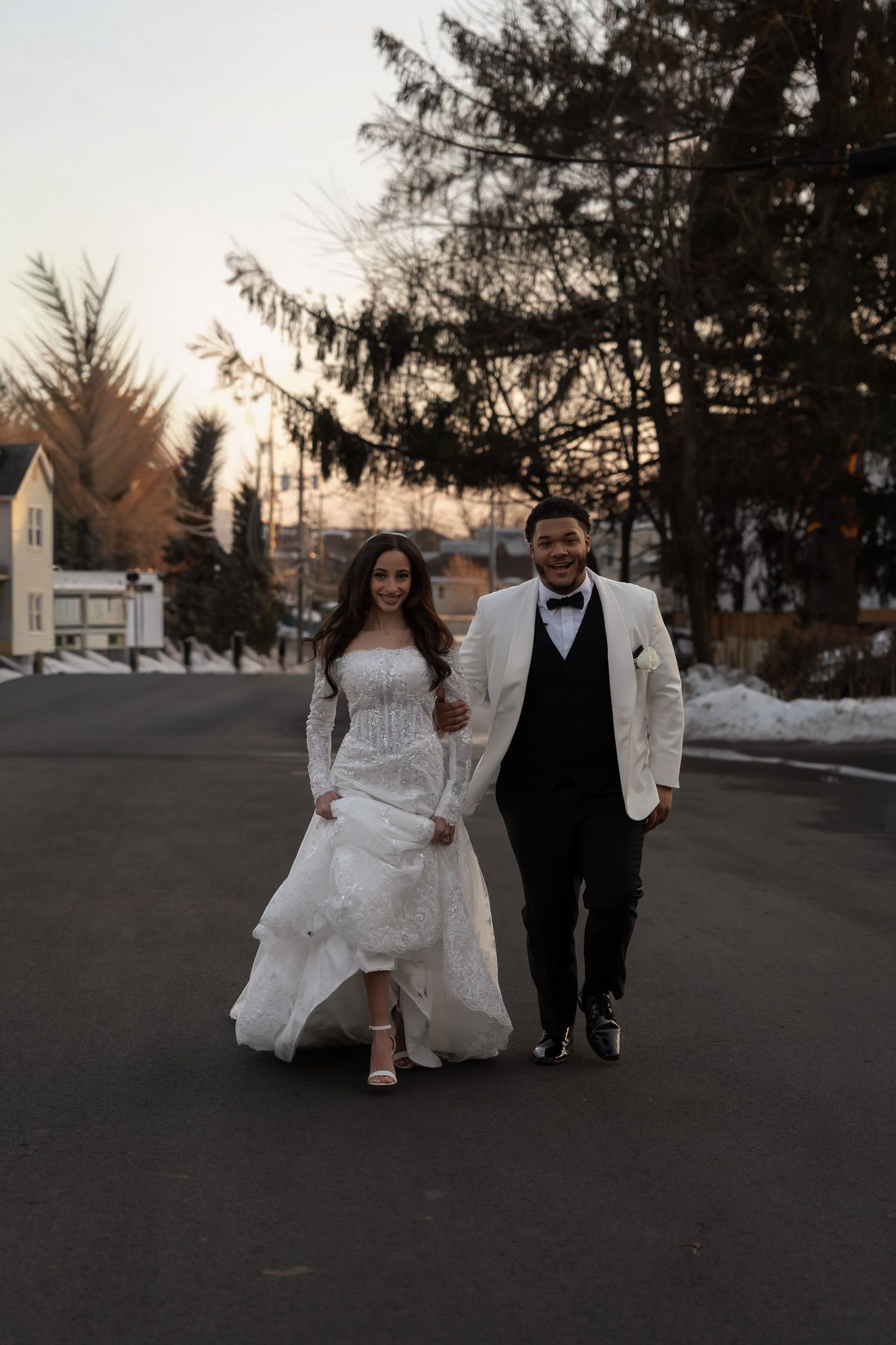 A couple in wedding attire walks down a snowy street at dusk with evergreen trees in the background.