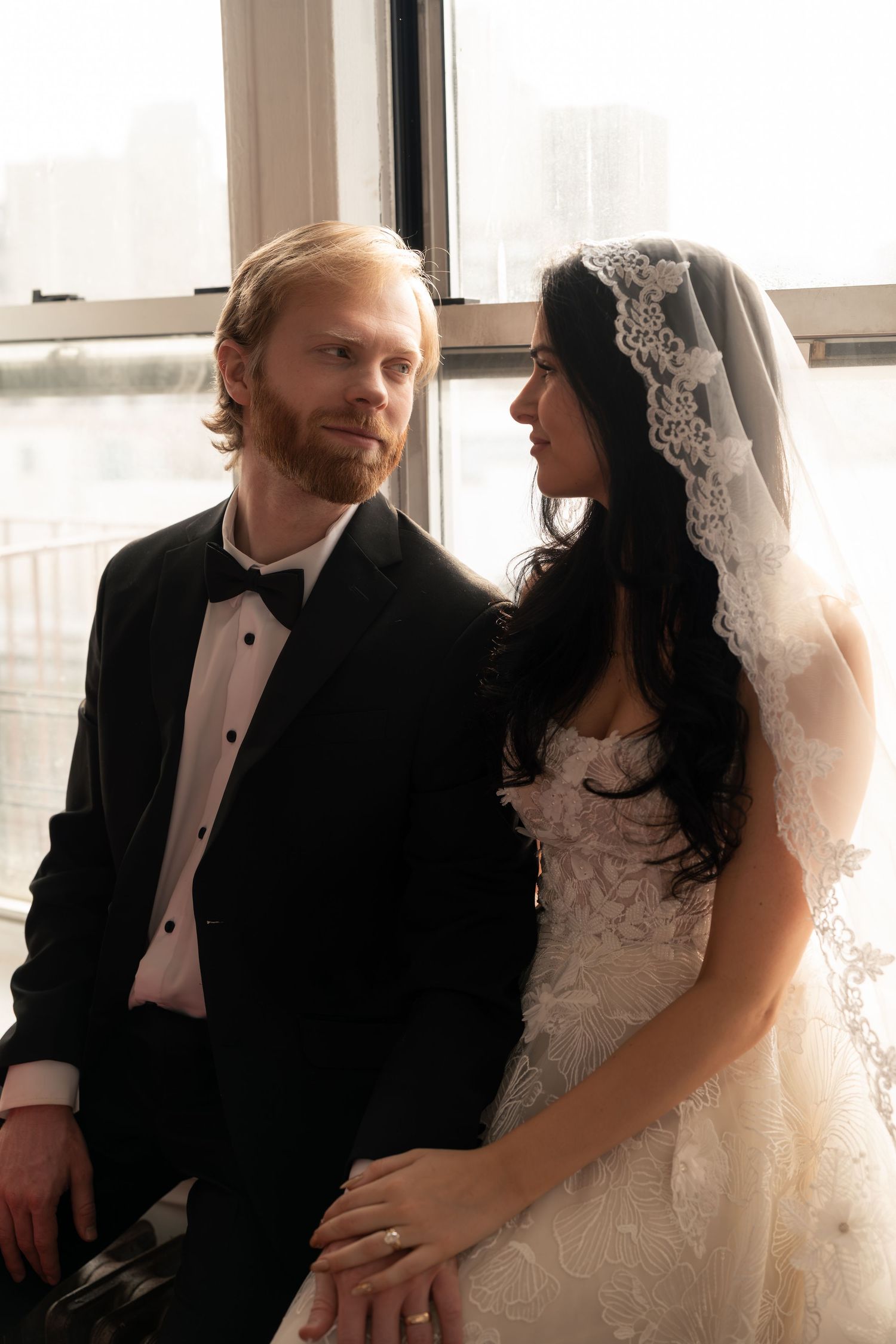 A bride and groom share intimate moments during their wedding photoshoot in front of large windows.