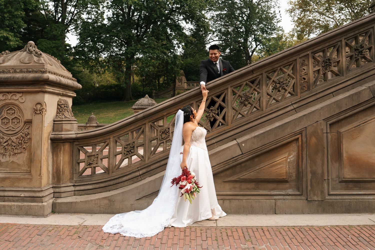 Bride in flowing white wedding dress poses gracefully on ornate stone steps surrounded by lush greenery in a garden setting.