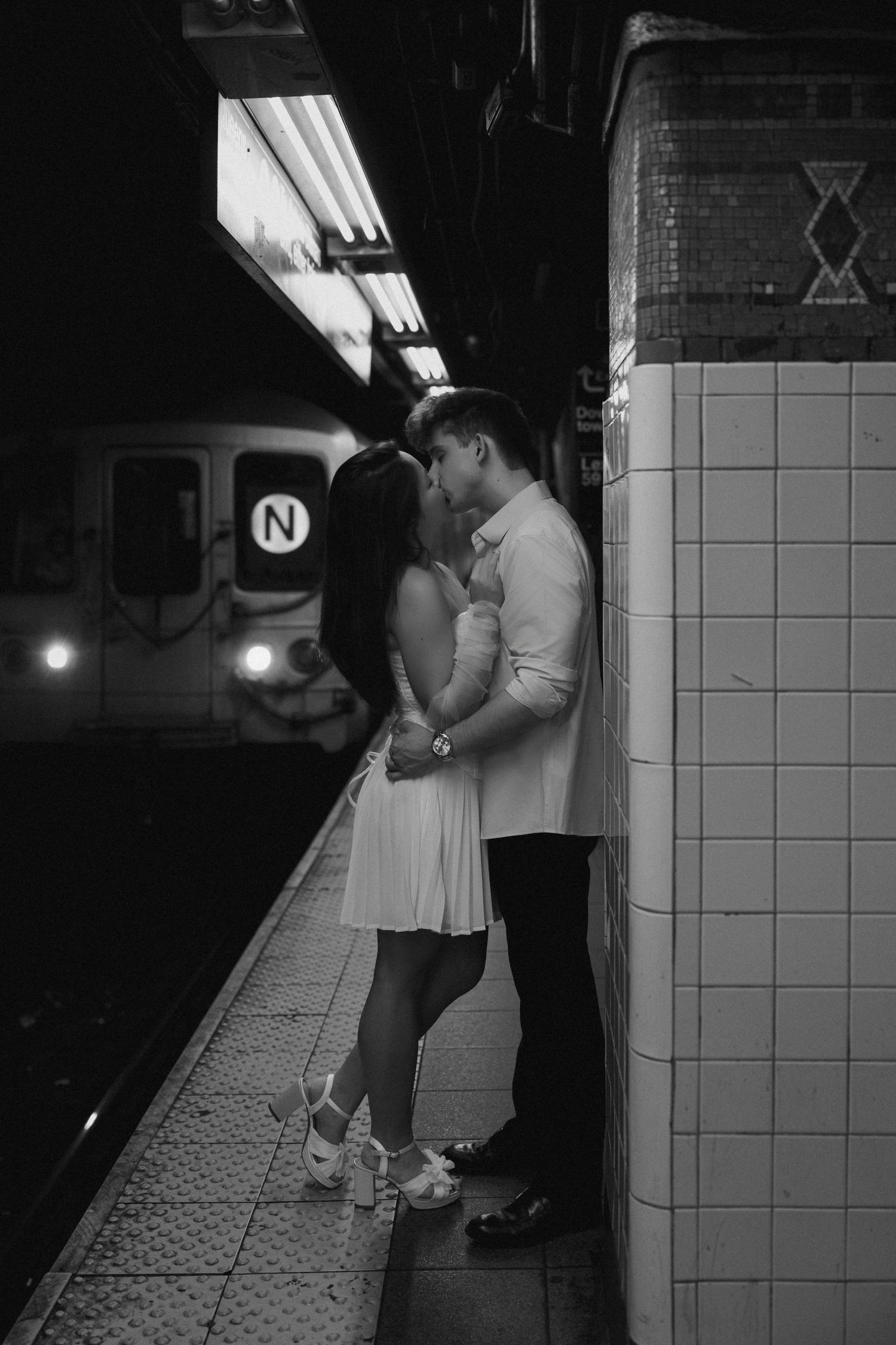 Couple kissing on NYC subway platform in black and white, N train approaching behind them.