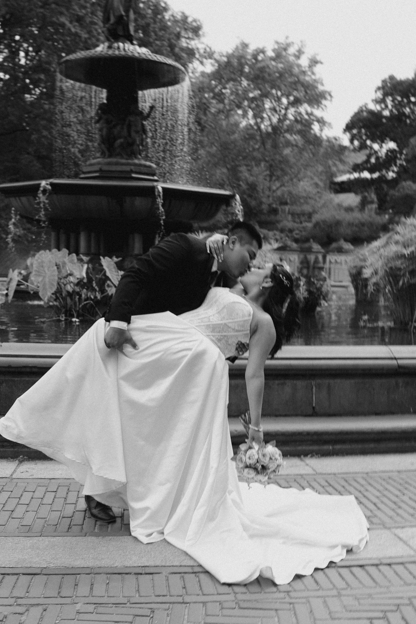 Black and white photo of newlyweds in dramatic poses by a fountain.