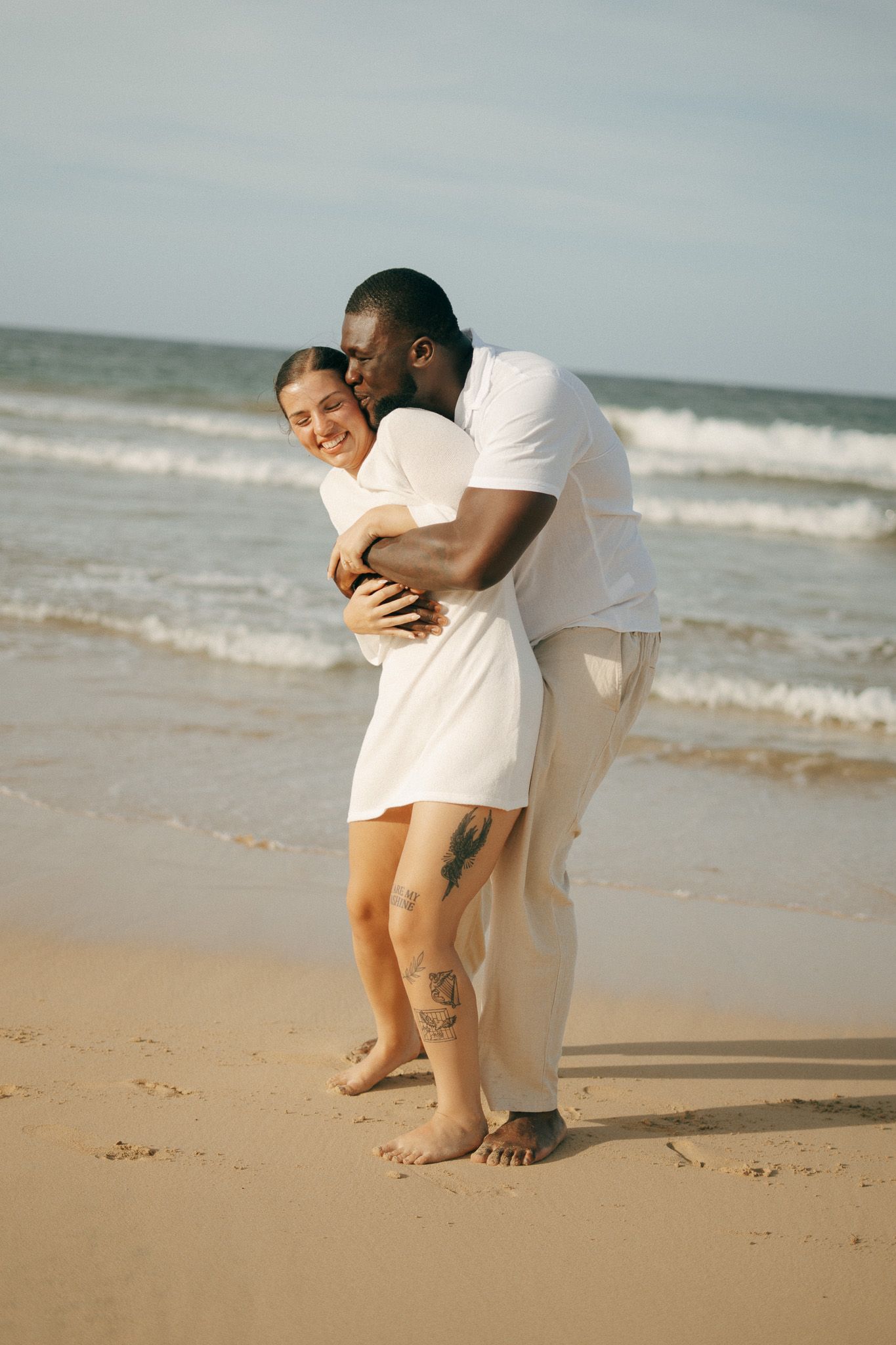 Happy interracial couple hugging barefoot on sandy beach, both wearing white outfits with ocean waves behind them.