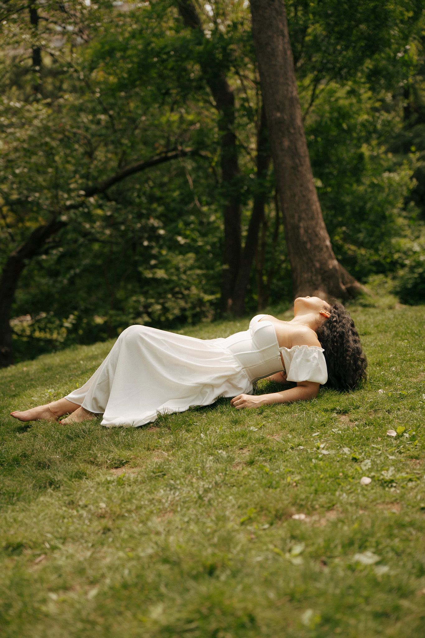 Woman in white off-shoulder dress lying on green grass in a lush forest setting.