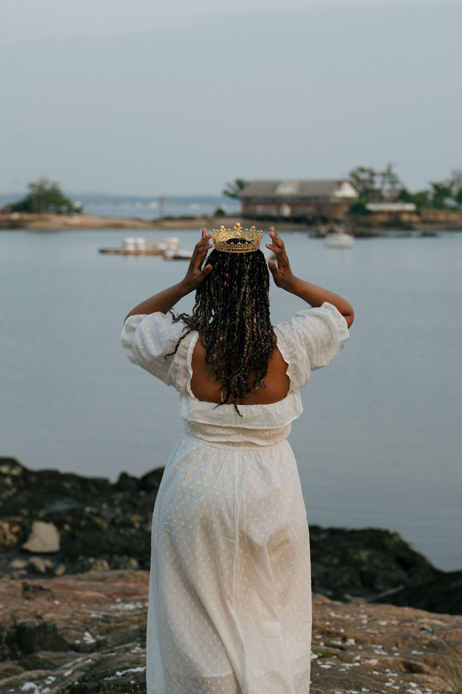 Person in white dress posing by the ocean with arms outstretched.